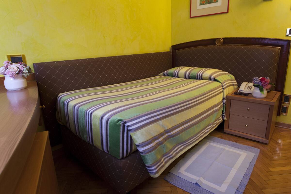 A hotel room with a bed, a bedside table with a phone and a vase of flowers, and a wooden dresser.