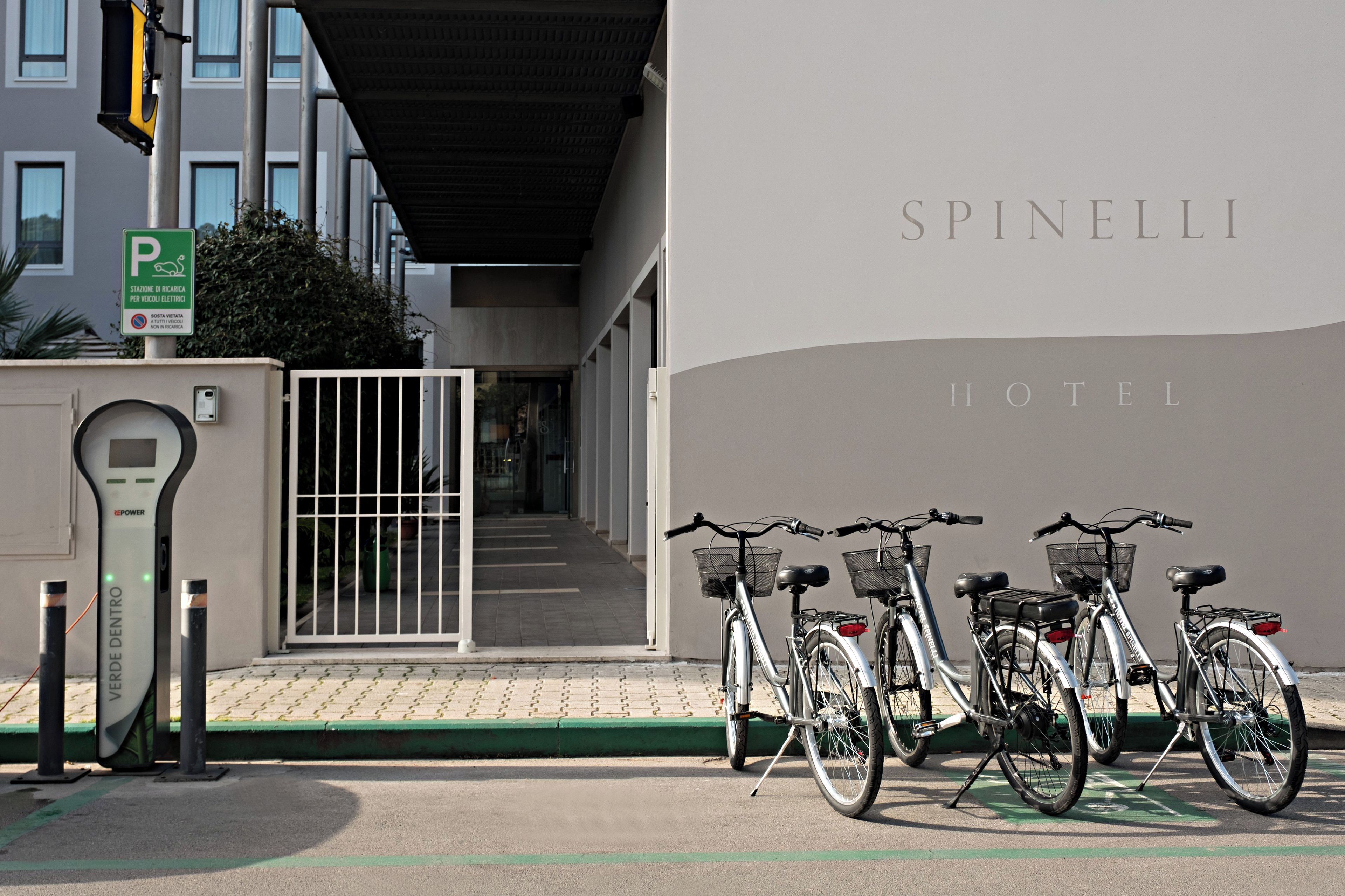 Entrada de un hotel con zona de estacionamiento para bicicletas y un letrero que indica la disponibilidad de carga para vehículos eléctricos.