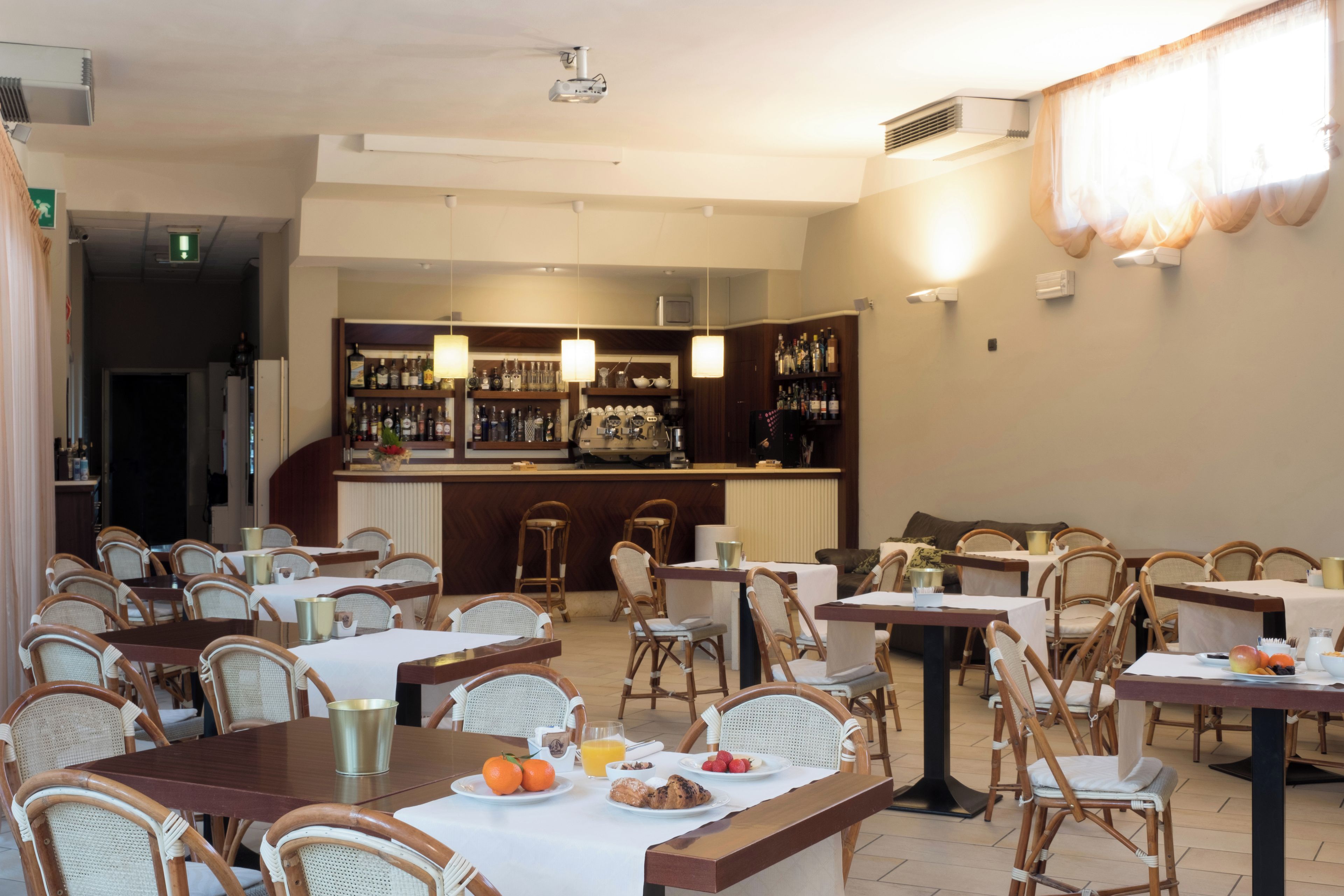 A restaurant with multiple tables and chairs, a bar area with shelves of bottles, and a ceiling-mounted projector.