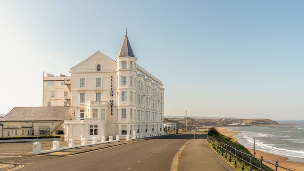 A coastal hotel with a prominent tower, a paved walkway along the beach, and a clear view of the ocean. - The Clifton Hotel (Scarborough)