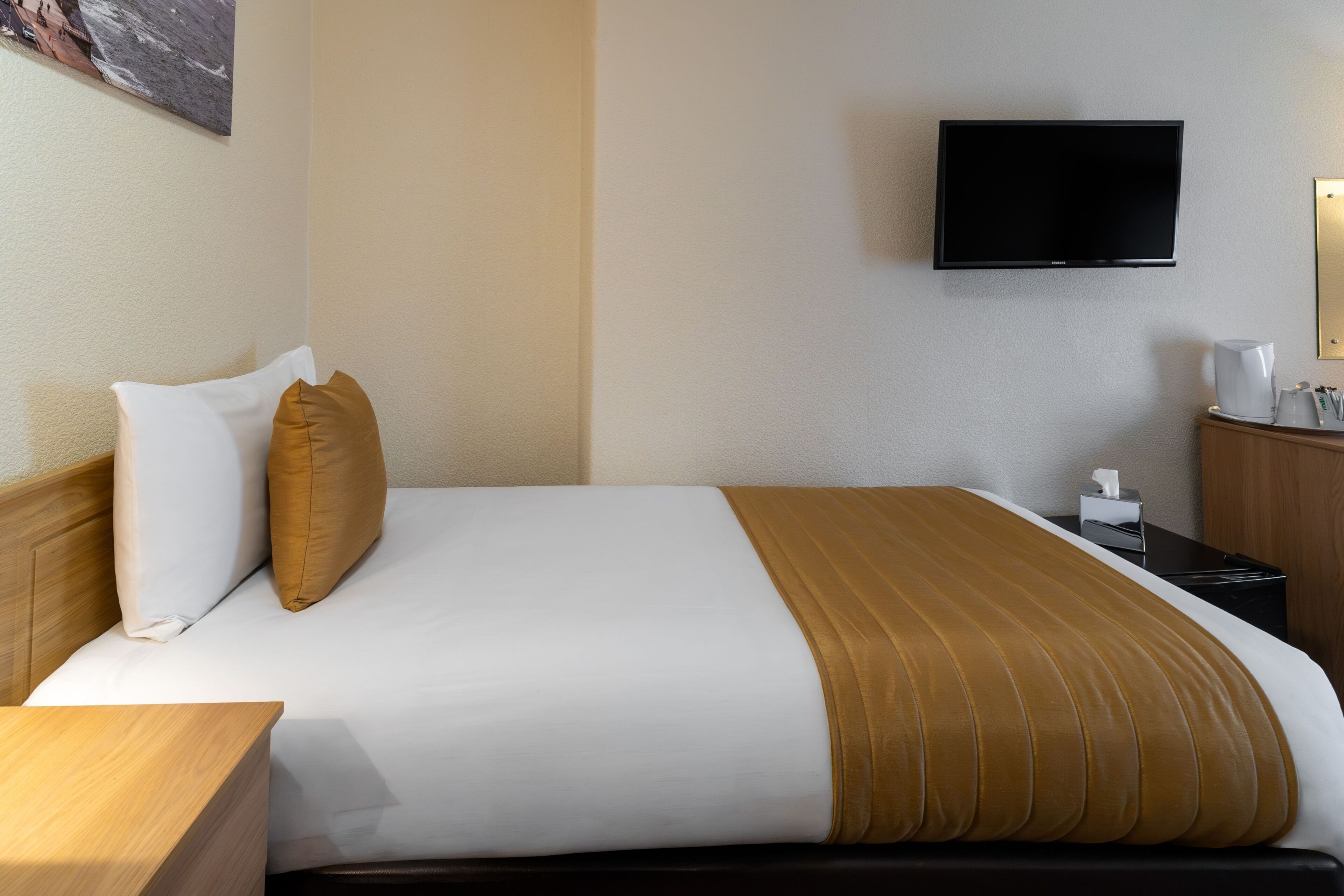 Une chambre d’hôtel comprenant un lit, un téléviseur fixé au mur, une table de chevet avec une lampe et une petite table de nuit avec une machine à café.