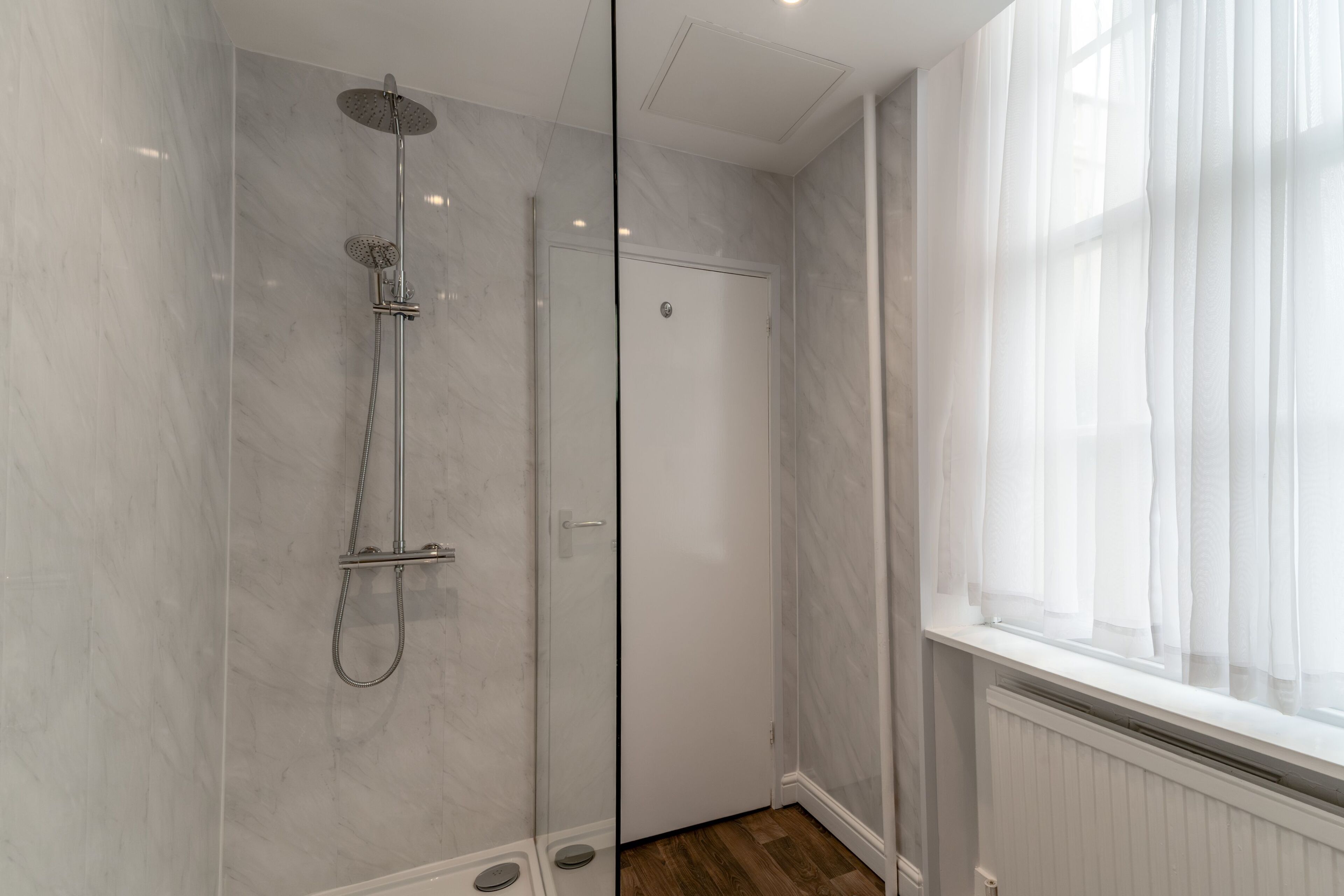 Een moderne badkamer met een douchecabine van glas, een witte deur en een raam met dunne gordijnen.