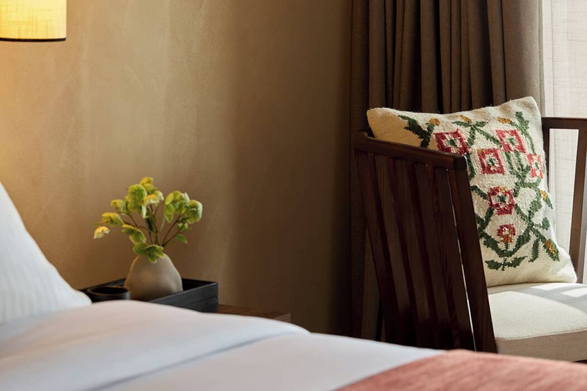 Hotellihuone, jossa on sänky, yöpöytä lampun kanssa, pehmustettu tuoli ja seinälle kiinnitetty peili.
