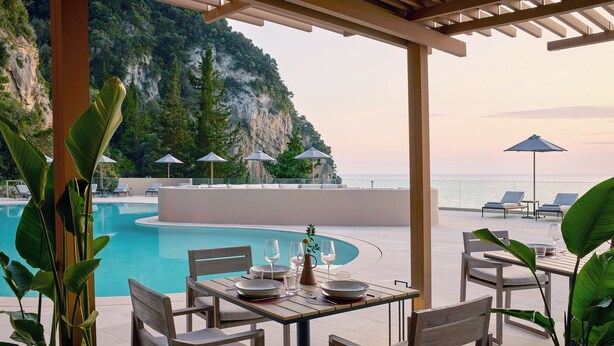 A poolside dining area with a table set for two, surrounded by chairs, and overlooking a pool with a view of the sea and a cliff. - Ella Alkyna – Adults Only (Corfu)