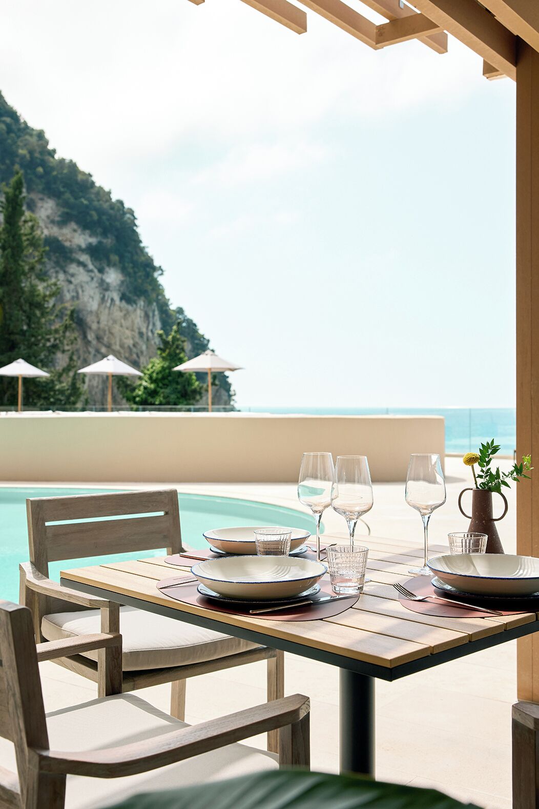 A wooden table set for four with plates, glasses, and cutlery, overlooking a pool and a mountainous landscape.