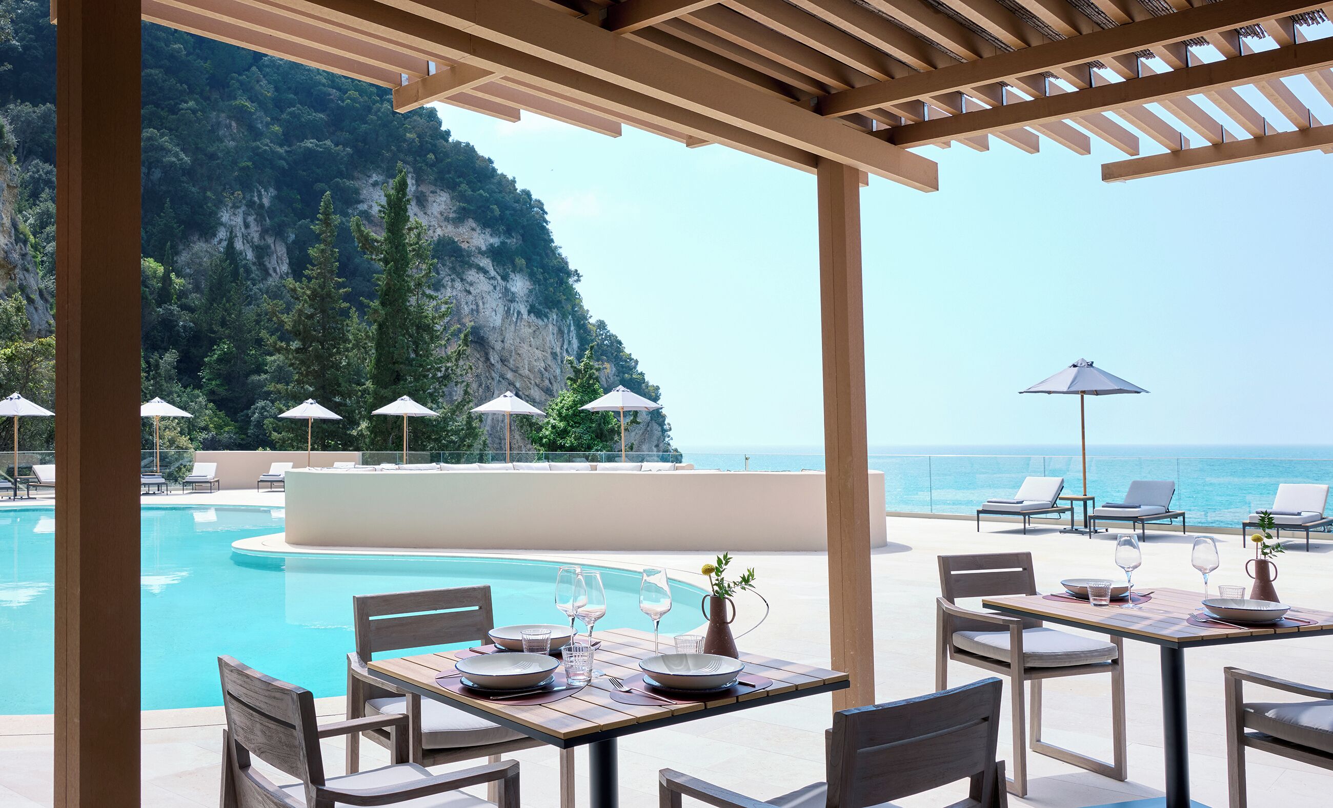 A poolside area with lounge chairs, umbrellas, and a table set for dining, overlooking a mountainous coastline.