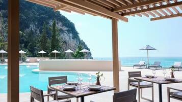 A poolside area with lounge chairs, umbrellas, and a table set for dining, overlooking a mountainous coastline.