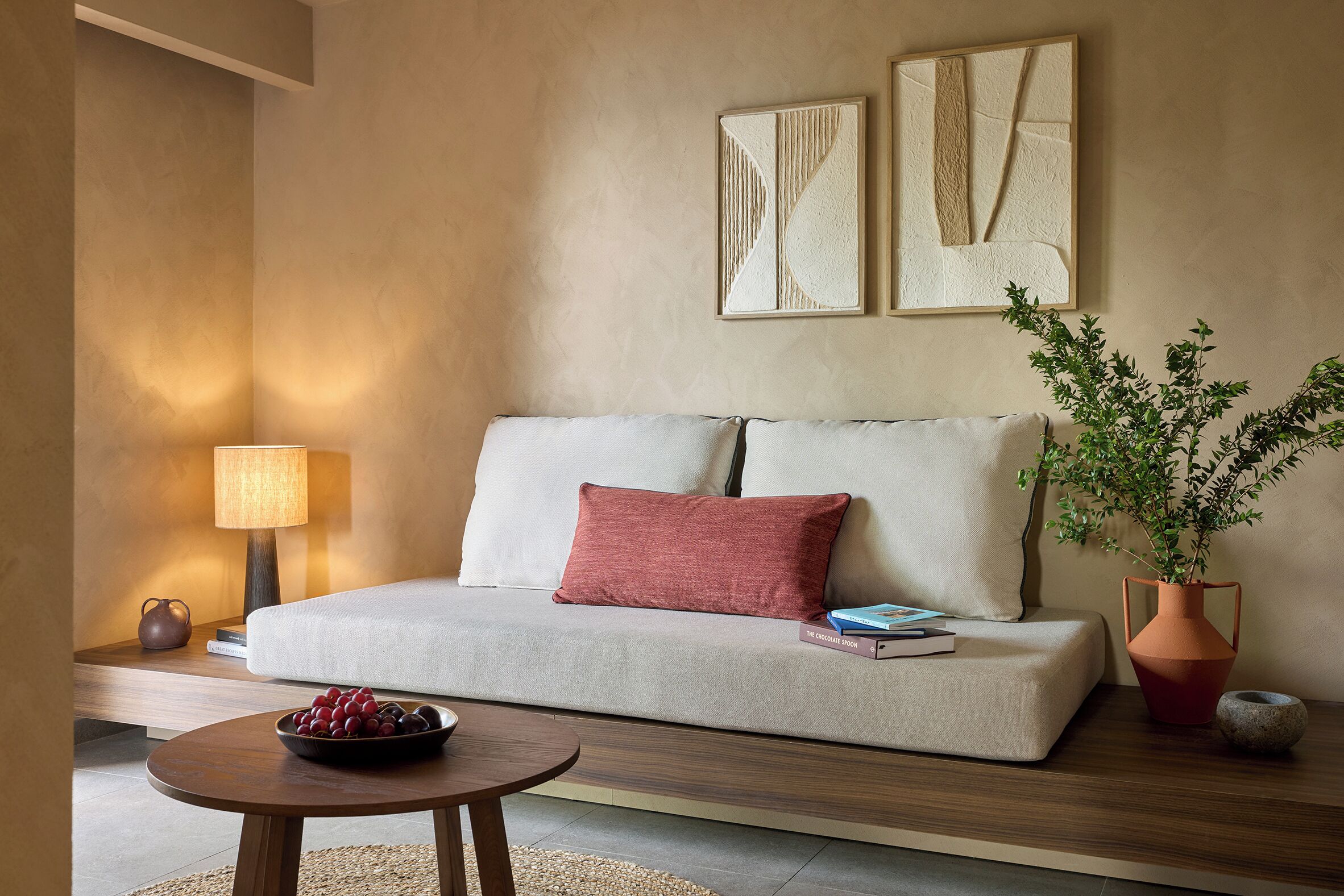 A modern living room with a sofa, a red pillow, a bowl of fruit, a lamp, and two abstract wall art pieces.