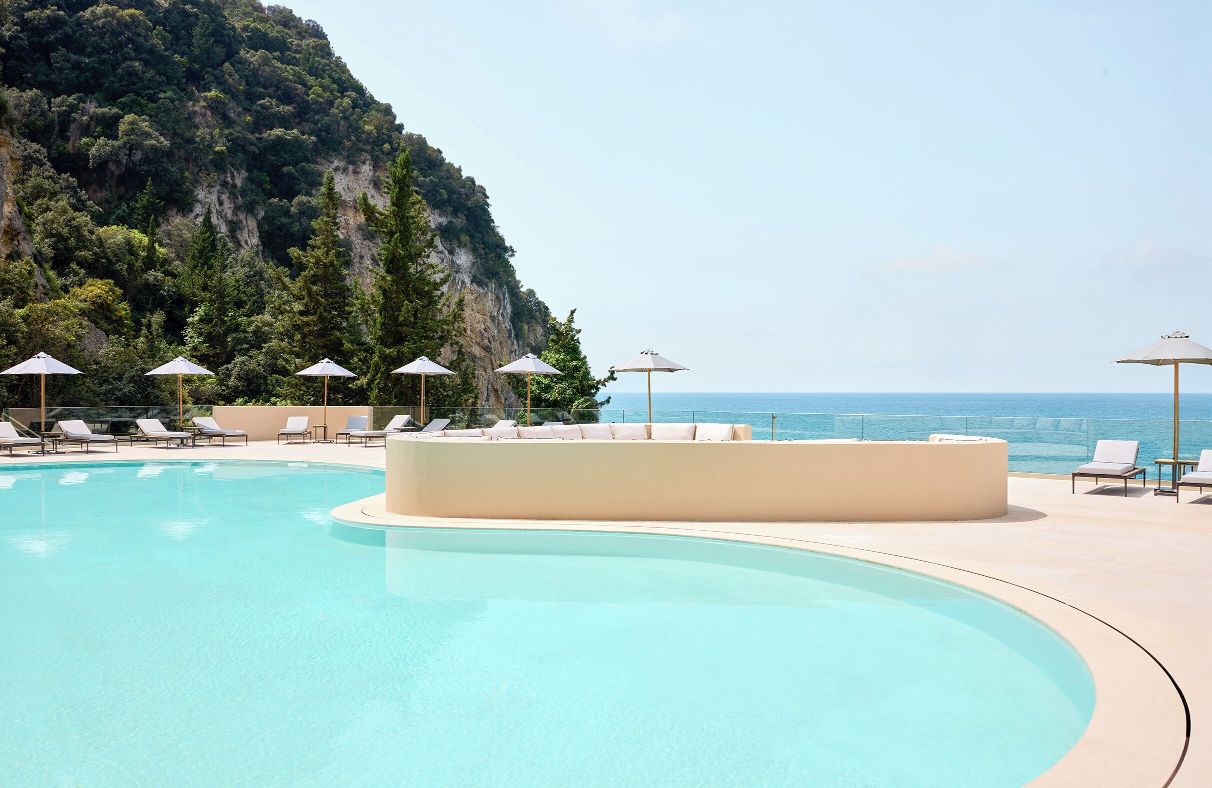 A pool with lounge chairs and umbrellas, surrounded by a cliff and the sea.