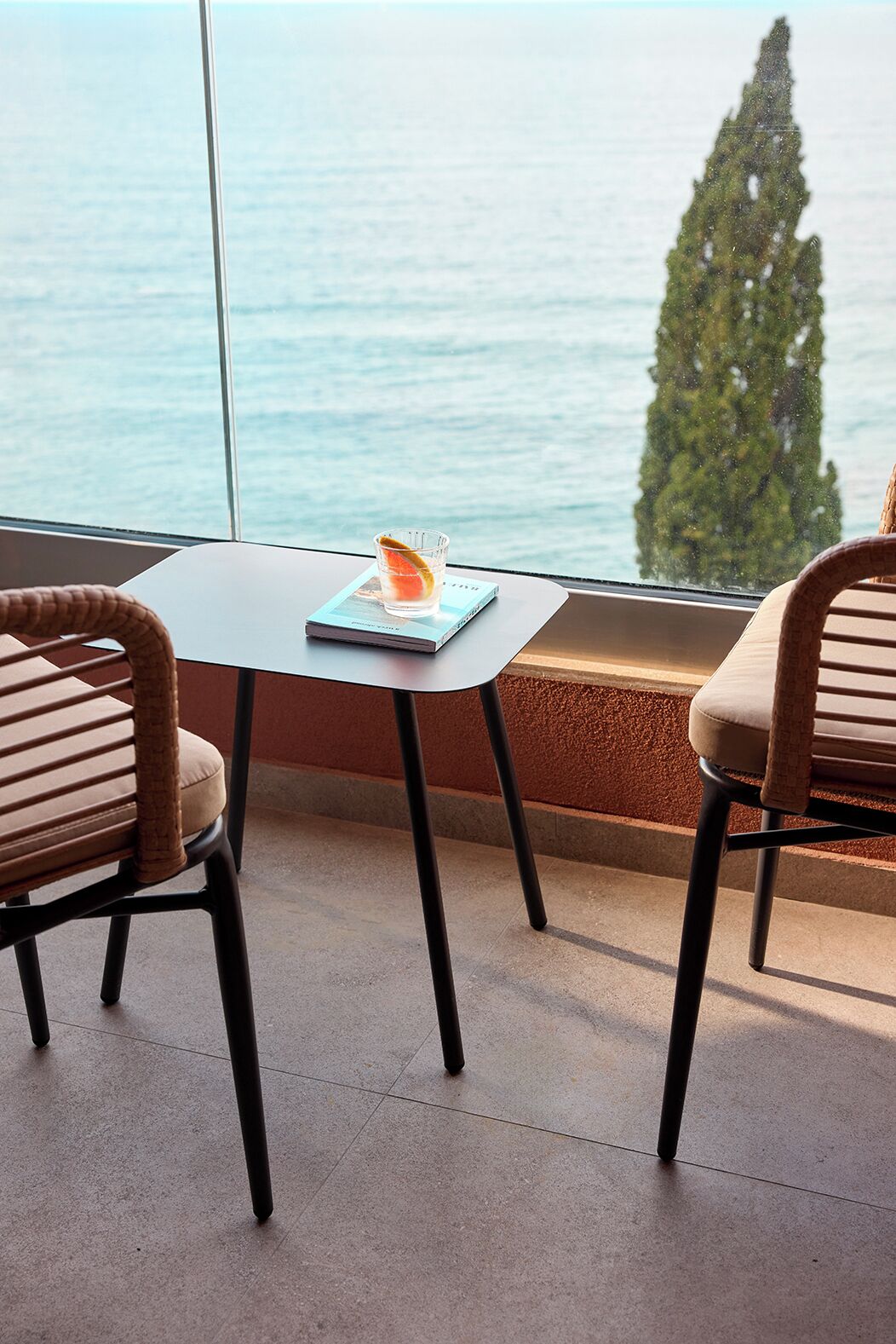 A modern room with a view of the sea, featuring two wicker chairs, a small table with a book and a glass of water, and a large window.