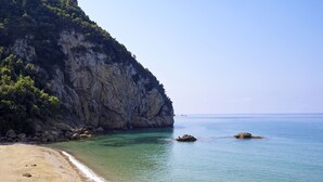 A beach with clear water, rocky cliffs, and a couple walking along the shore. - Ella Alkyna – Adults Only (Corfu)