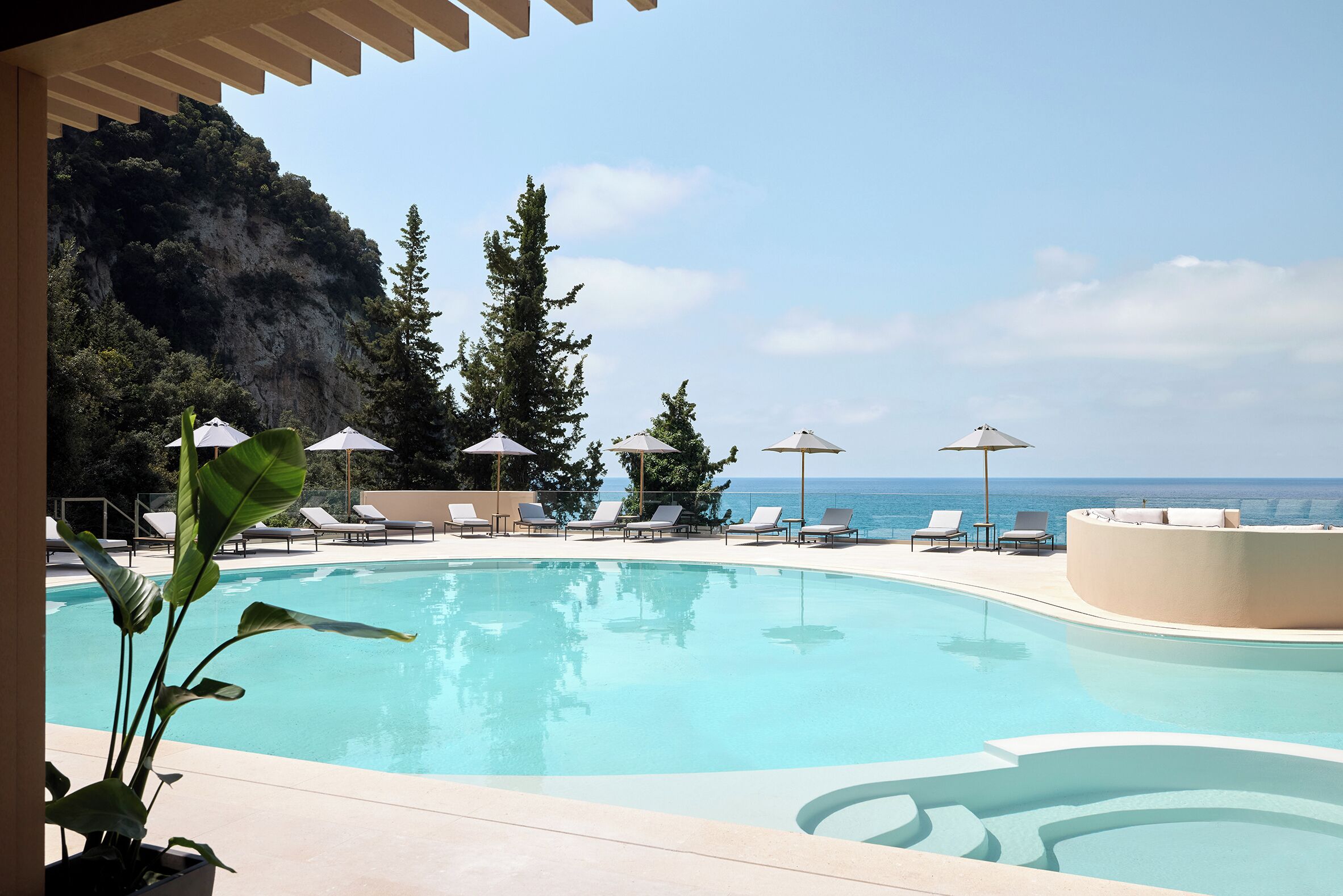 A swimming pool with lounge chairs and umbrellas, surrounded by trees and overlooking the ocean.