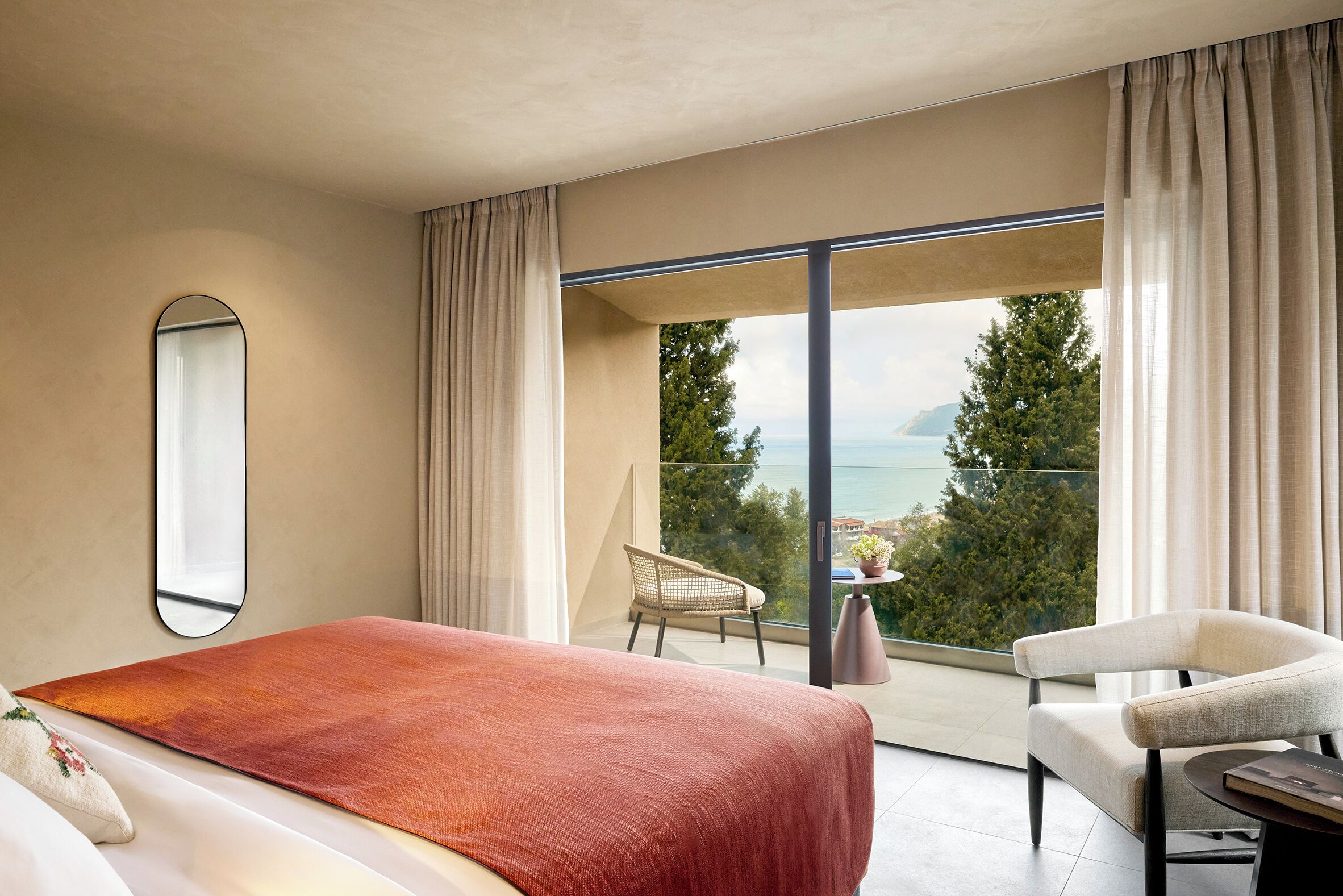 A modern hotel room with a large bed, a chair, a small table, and a balcony with a view of the sea.