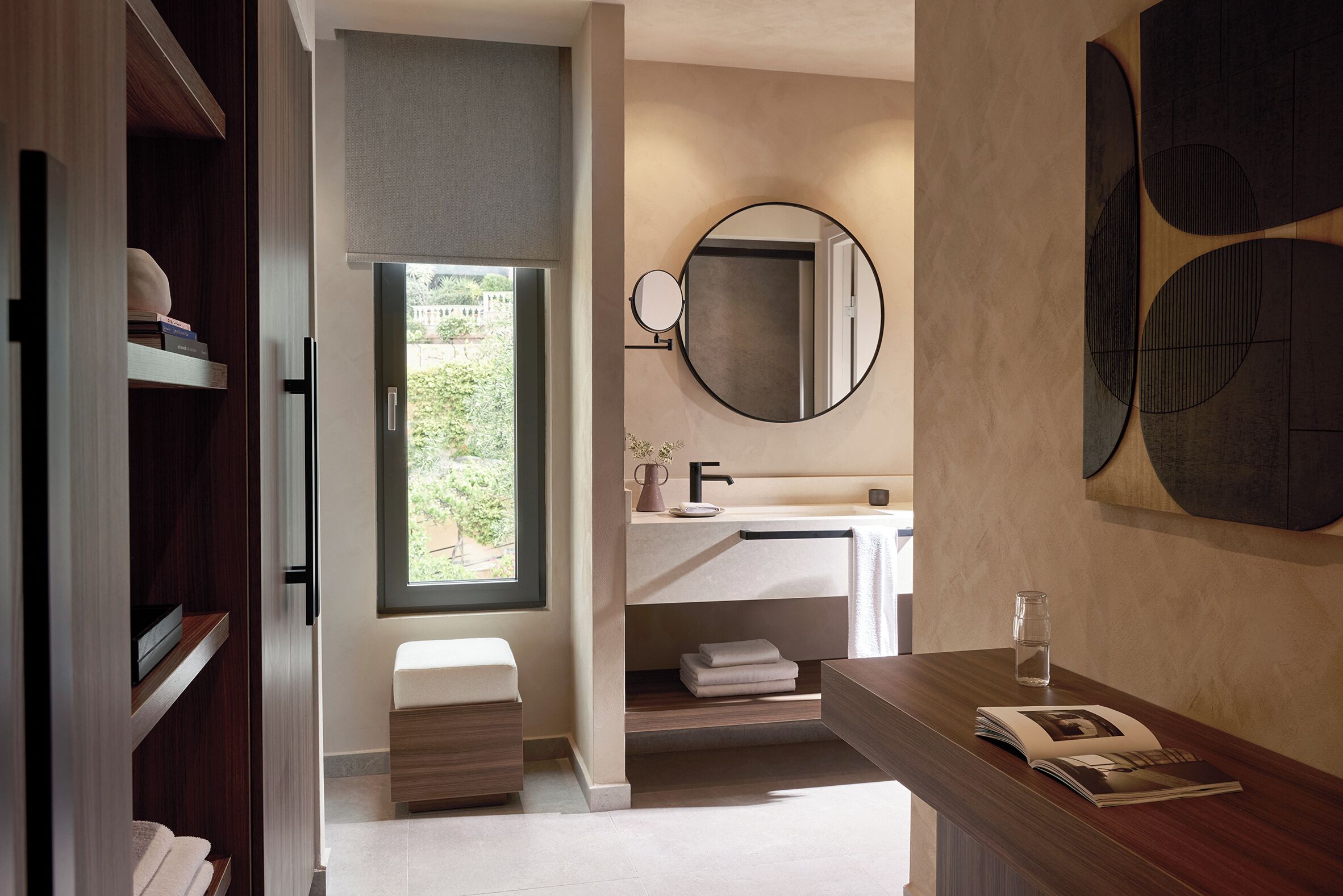Une salle de bain moderne avec un lavabo intégré, un miroir rond et une fenêtre donnant sur un espace verdoyant.
