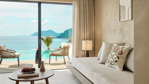 A hotel room with a large window overlooking a pool and mountains. There is a sofa, two chairs, a table with a book and a pitcher, and a lamp. - Ella Alkyna – Adults Only (Corfu)