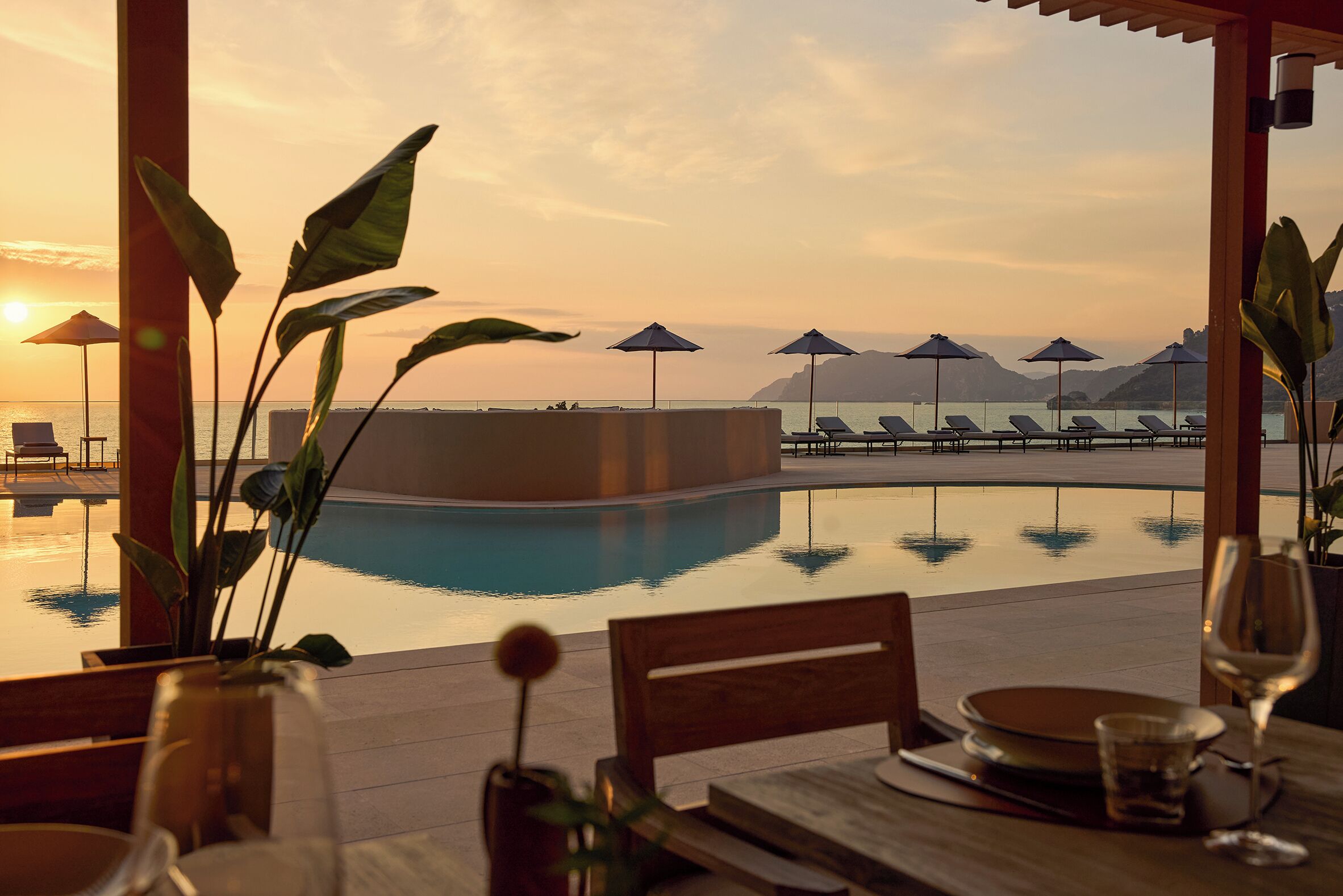 A poolside dining area with a view of the ocean and mountains at sunset.