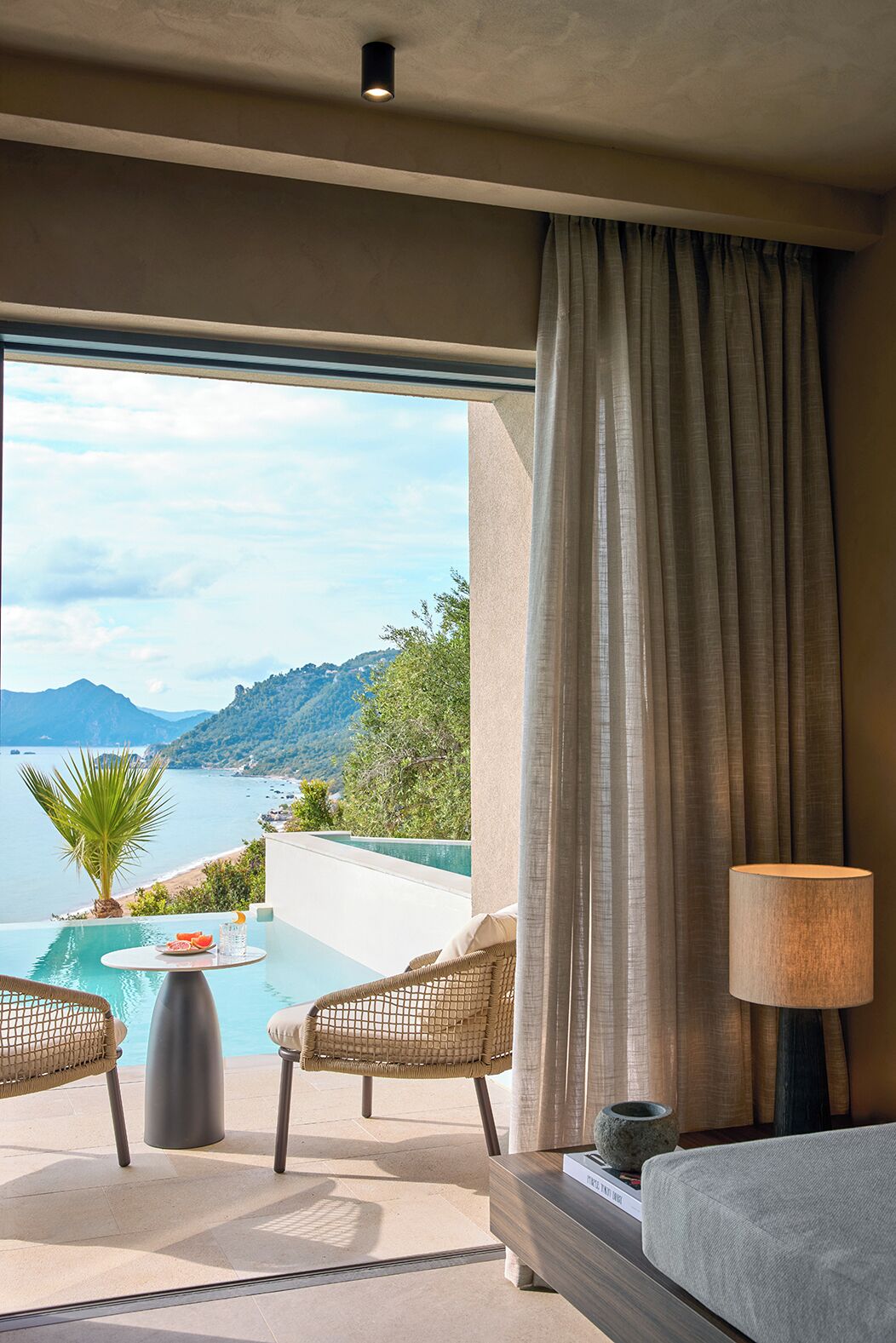A room with a view of a beach and mountains, featuring a pool, two wicker chairs, a table with fruit, and a lamp.