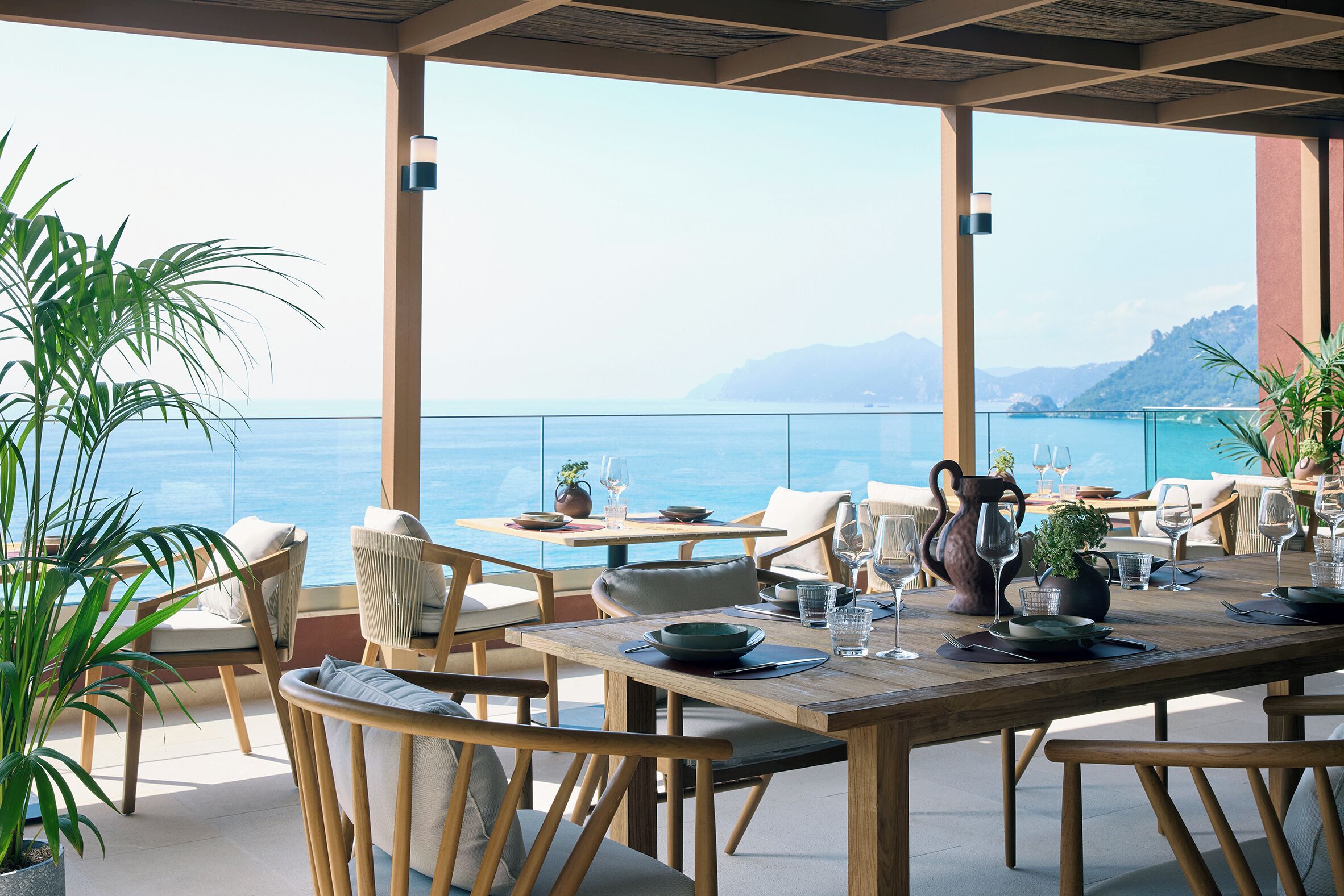 A terrace with wooden tables and chairs, potted plants, and a view of the sea.