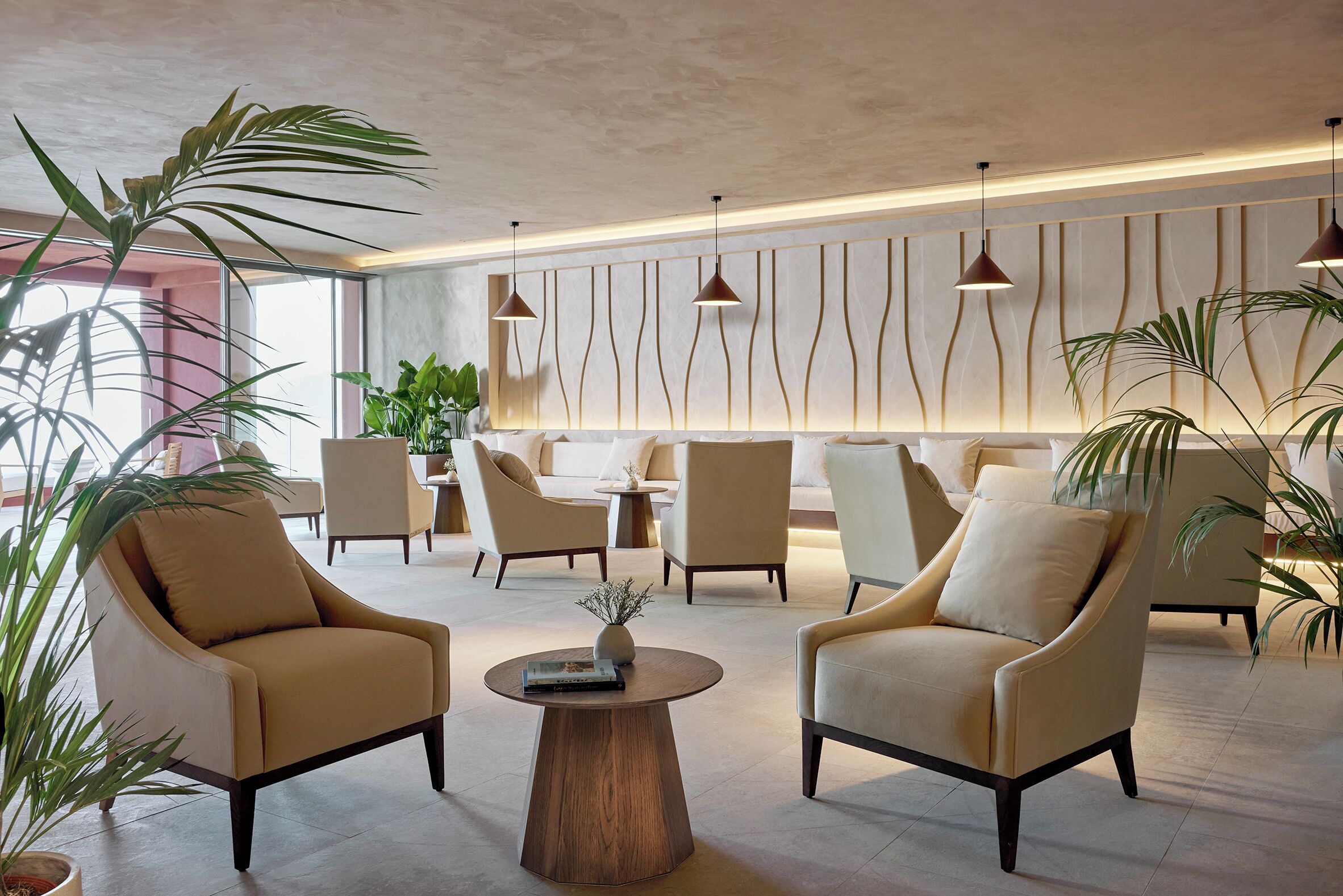 Un hall d’entrée moderne avec deux fauteuils placés face à une table basse ronde en bois, un plafond haut avec des luminaires suspendus et une grande plante au premier plan.