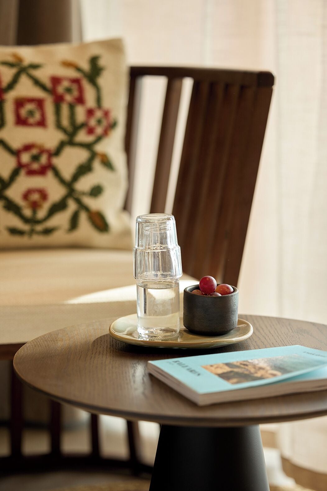 A round wooden table with a clear glass water bottle and a small bowl of fruit.