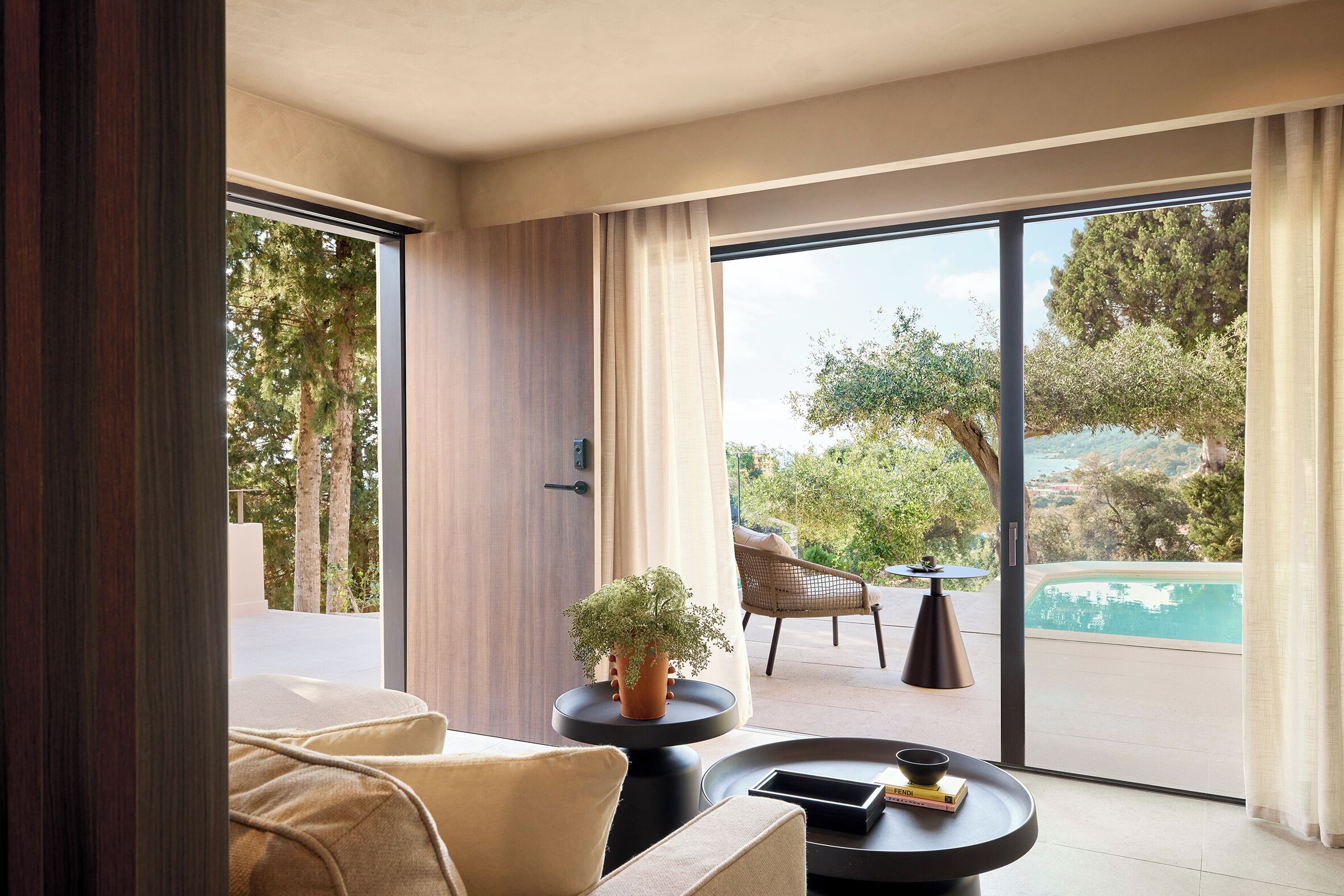 Una sala de estar moderna con un sofá, una mesa de centro y una planta en maceta. Hay un ventanal corredero que da a un área exterior con piscina y una silla.