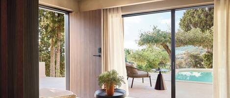 Uma sala de estar moderna com um sofá, uma mesa de centro e uma planta em vaso. Há uma porta de vidro deslizante que dá para uma área externa com piscina e uma cadeira.