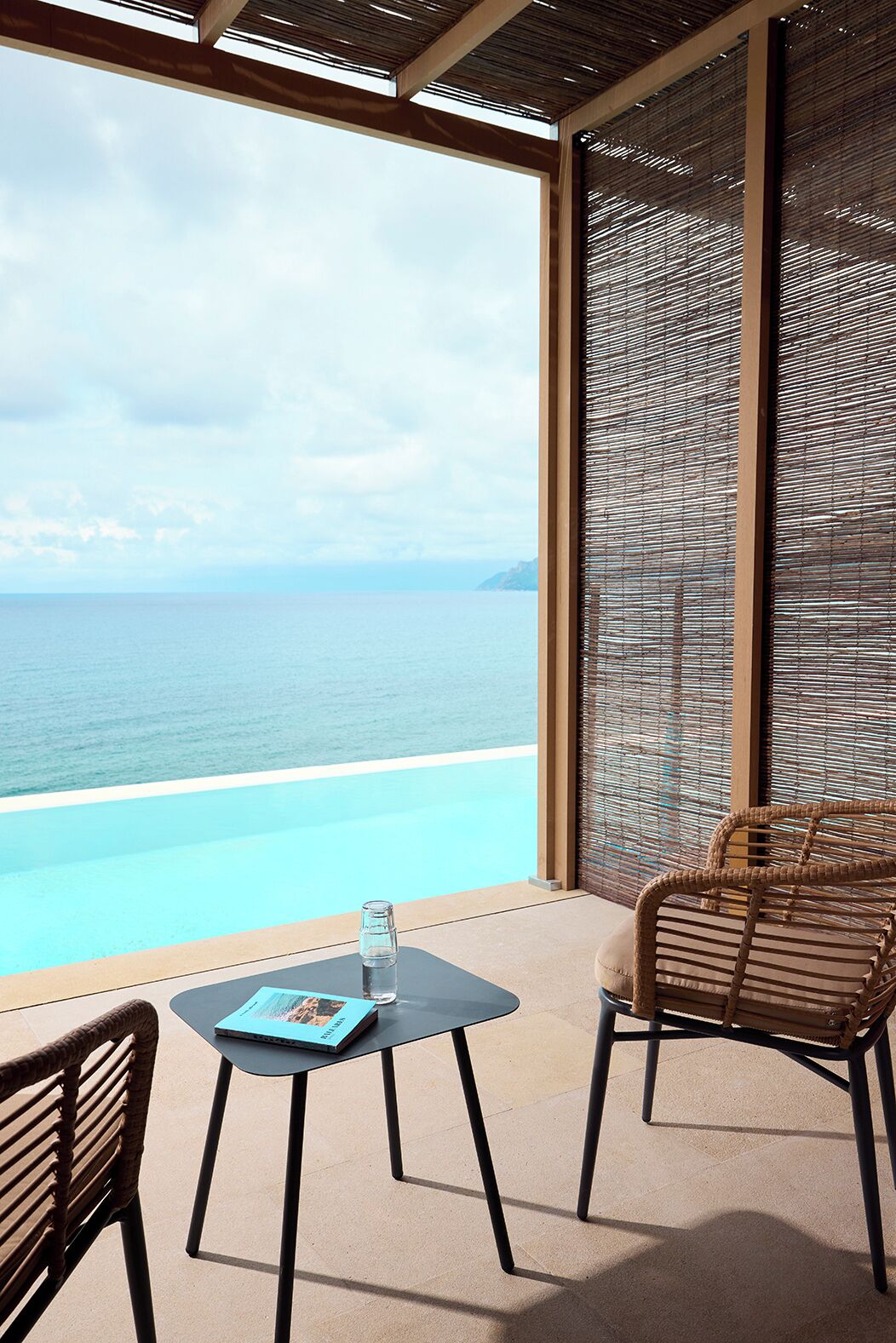 Un balcón con vista al mar, una mesa con un vaso de agua y sillas de mimbre.