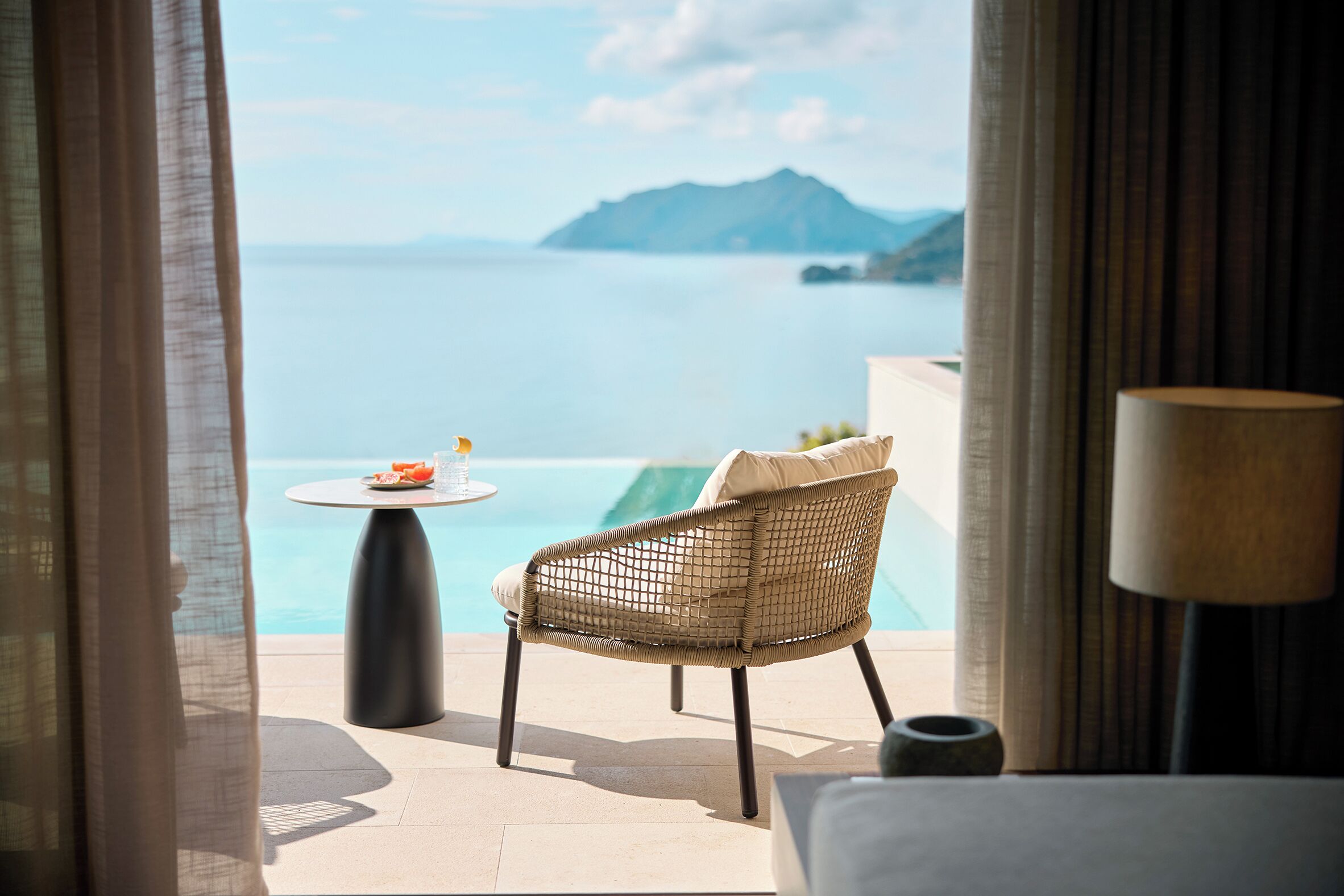 A room with a view of the sea, a wicker chair, a small table with a fruit bowl, and a lamp.