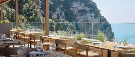 An outdoor dining area with tables and chairs, overlooking a coastal cliff and the sea.