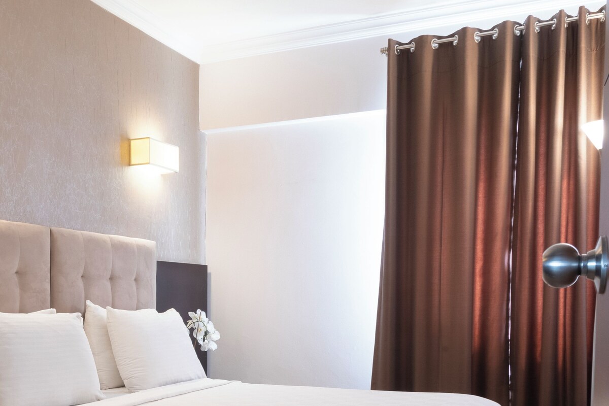 Habitación de hotel con una cama grande, cabecera, luz fijada a la pared, ventana con cortinas y puerta con manija.