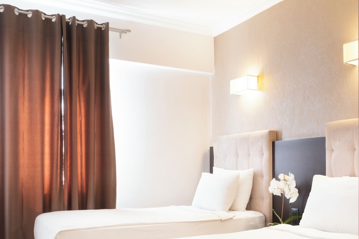 Habitación de hotel con dos camas, un ventanal con cortinas, una lámpara fijada a la pared y una alfombra estampada.