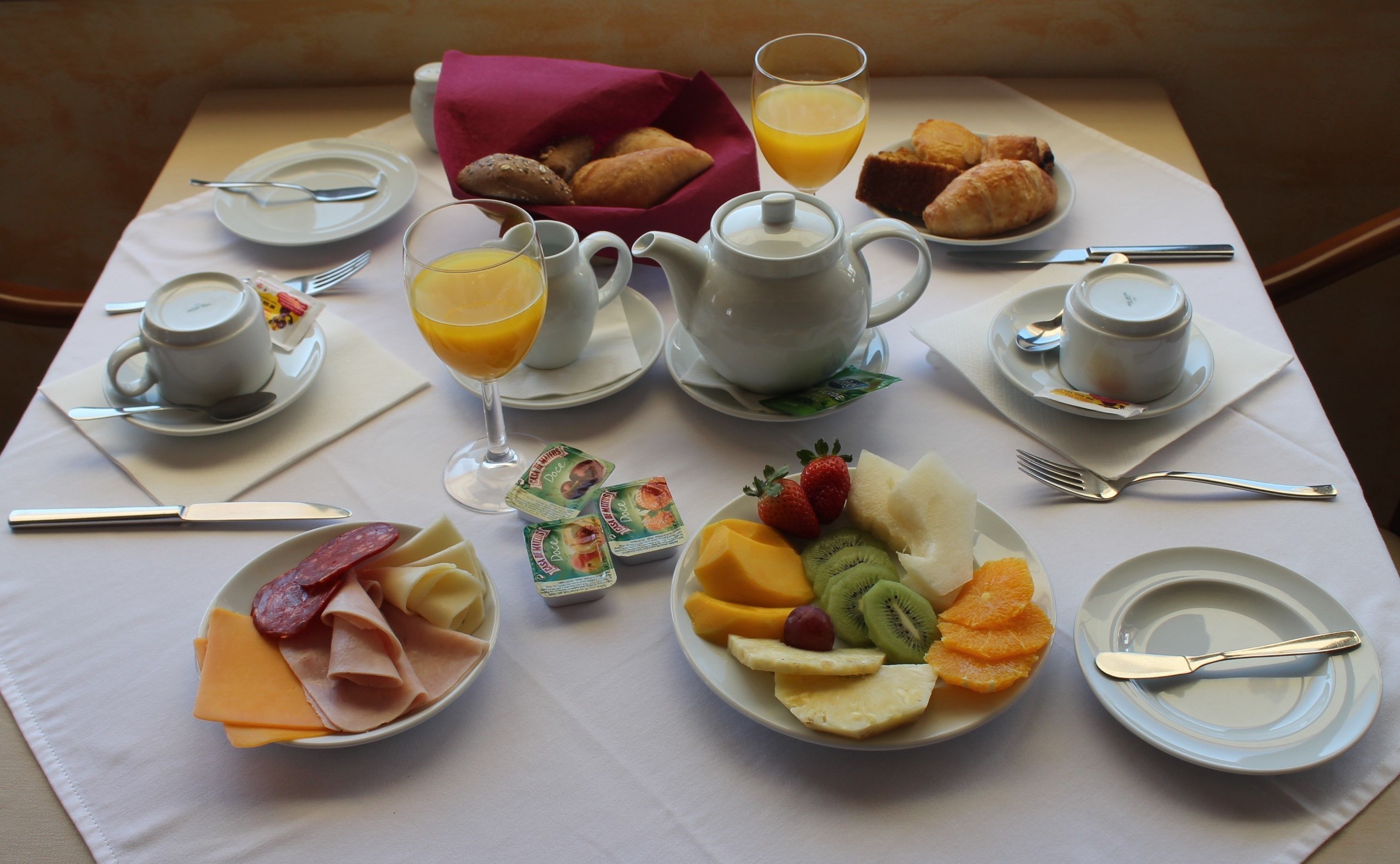 Un desayuno servido con platos de comida, una tetera, tazas y un vaso de zumo de naranja.