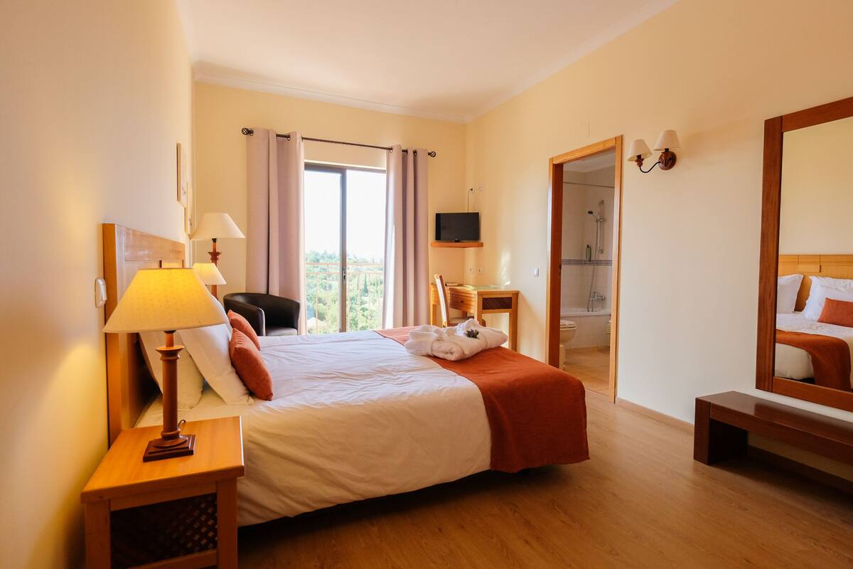 Een hotelkamer met een bed, nachtlampjes, een bureau, een stoel, een televisie, een douche en een grote spiegel.