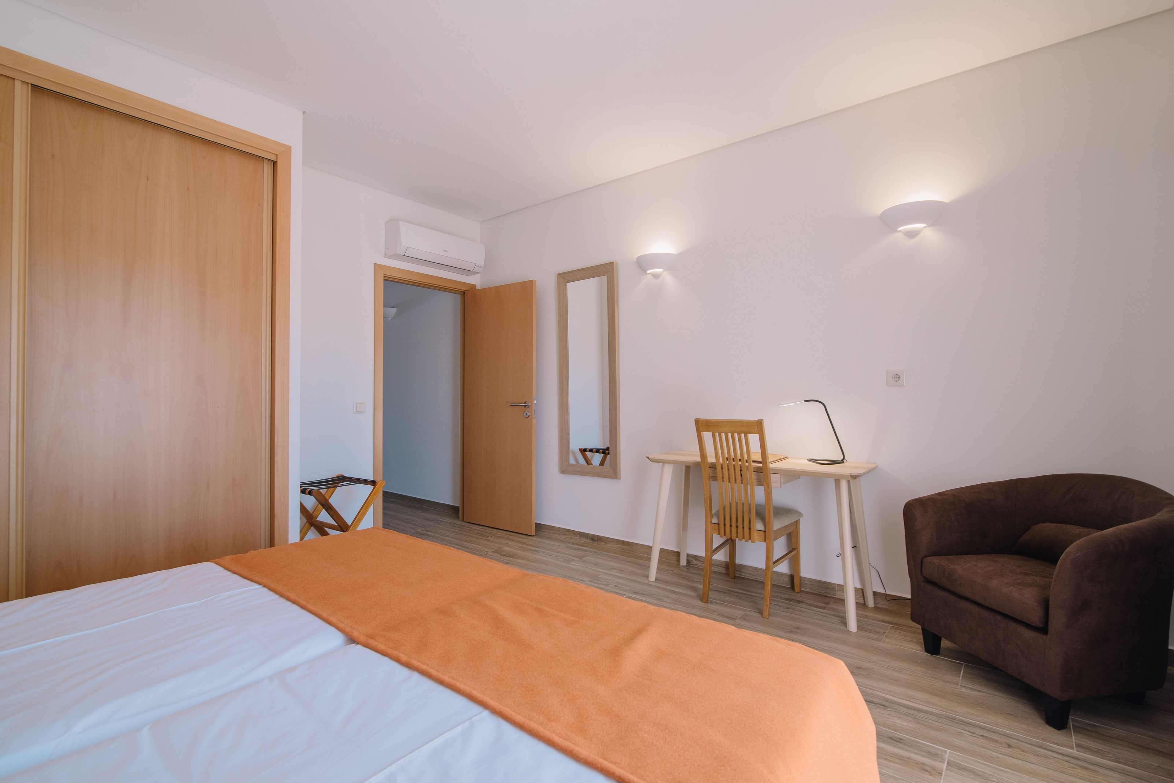 Una habitación de hotel con cama, escritorio con silla, sillón marrón y un espejo en la pared.