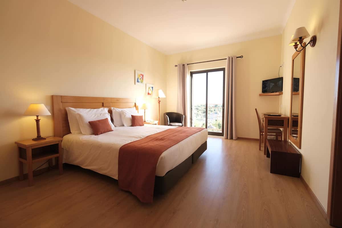 Een hotelkamer met een groot bed, nachtkastjes met lampen, een stoel, een bureau met stoel, en een groot raam met uitzicht.