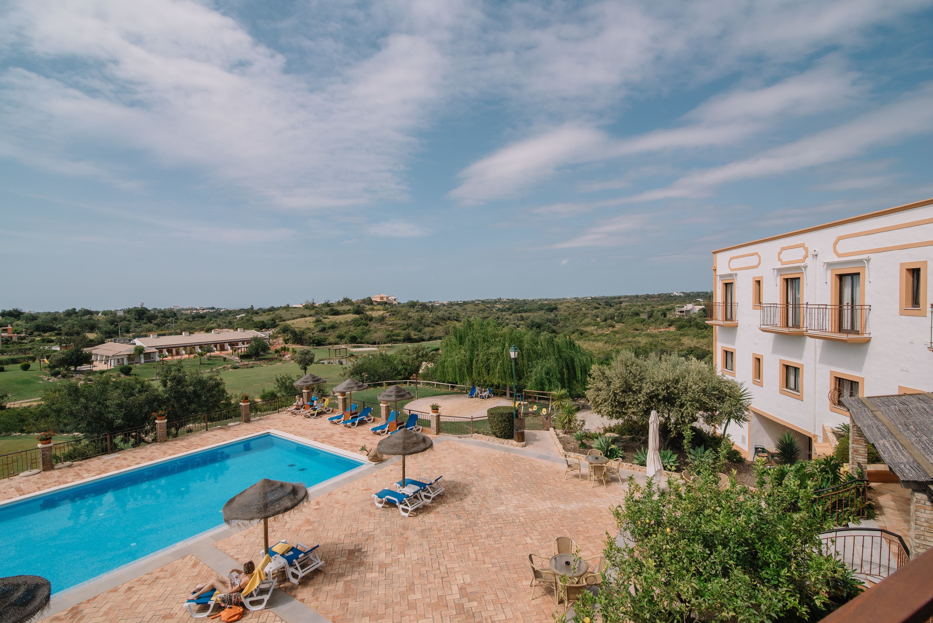 A pool area with lounge chairs and umbrellas, surrounded by greenery and a view of the countryside.