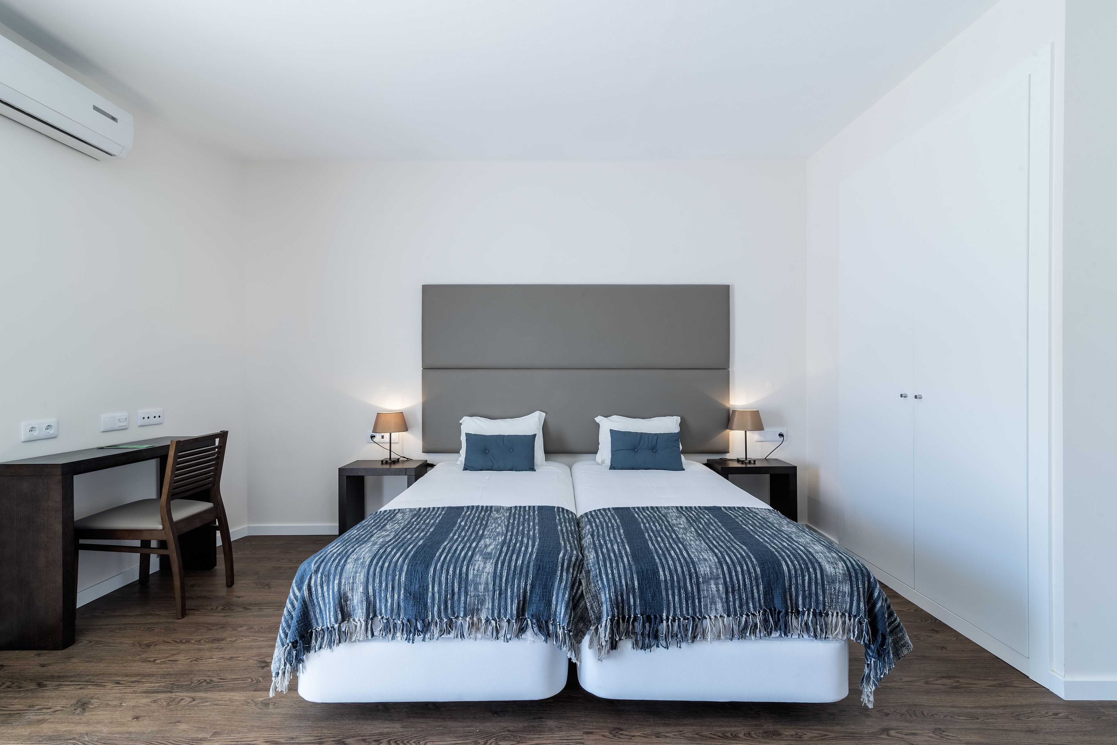 A double bed with a grey headboard, two bedside tables with lamps, a wooden desk, and a chair.