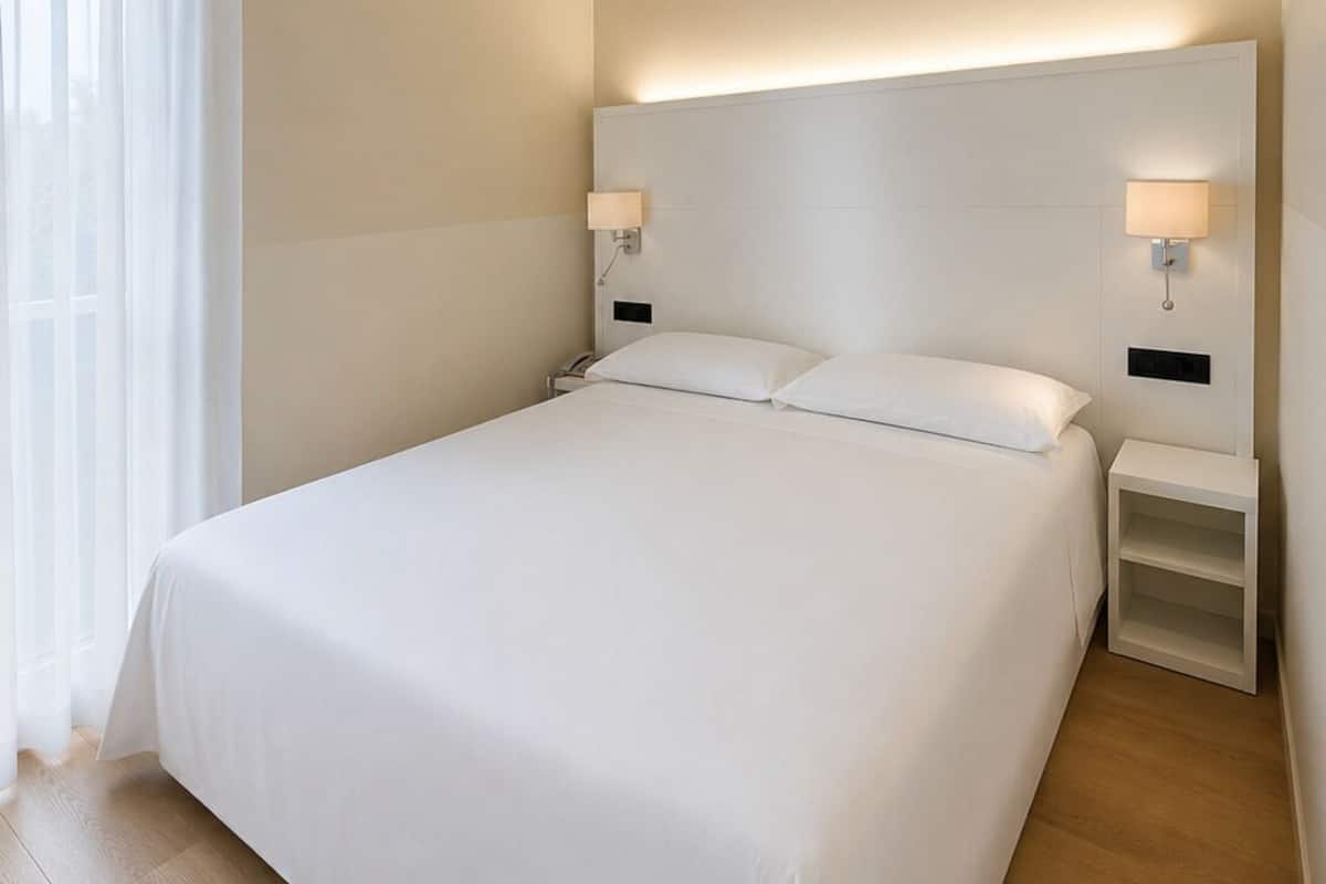 A hotel room with a large bed, two bedside tables with lamps, a window with curtains, and a wooden floor.