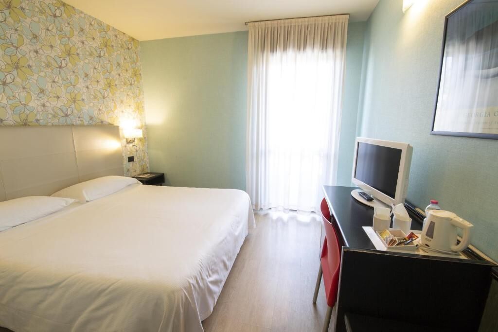 Ein Hotelzimmer mit einem Bett, einem Schreibtisch mit Computer, einem Stuhl und einem fenster mit Vorhängen.