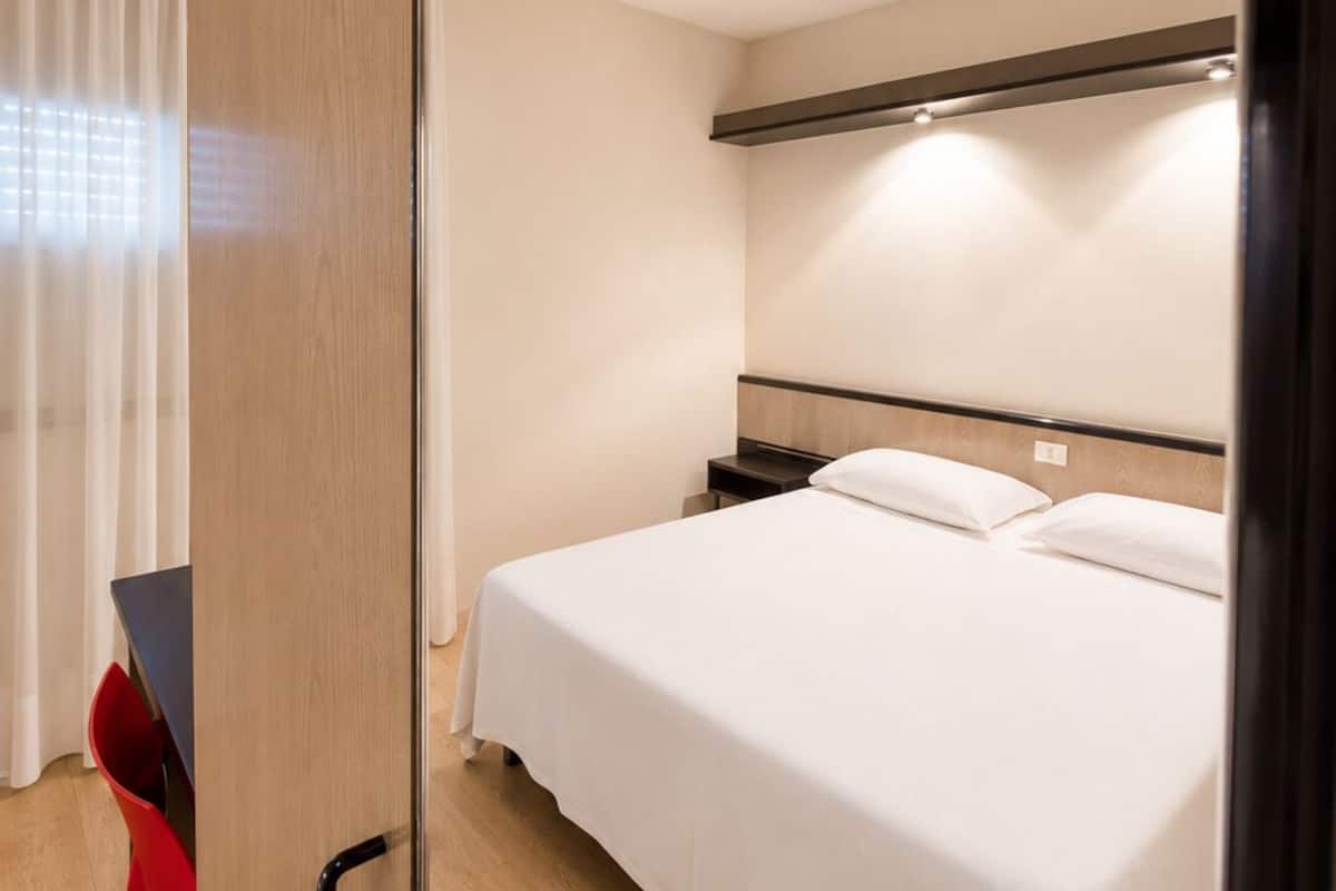 A hotel room with a bed, bedside tables, a desk with a chair, and a window with curtains.