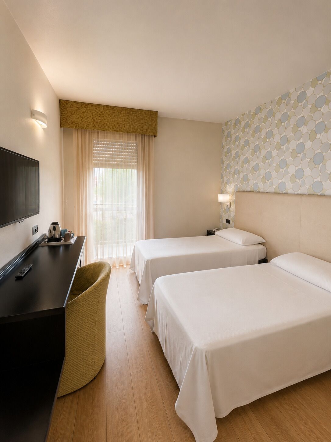 A hotel room with two beds, a desk with a television, a window with blinds, and a patterned wall.