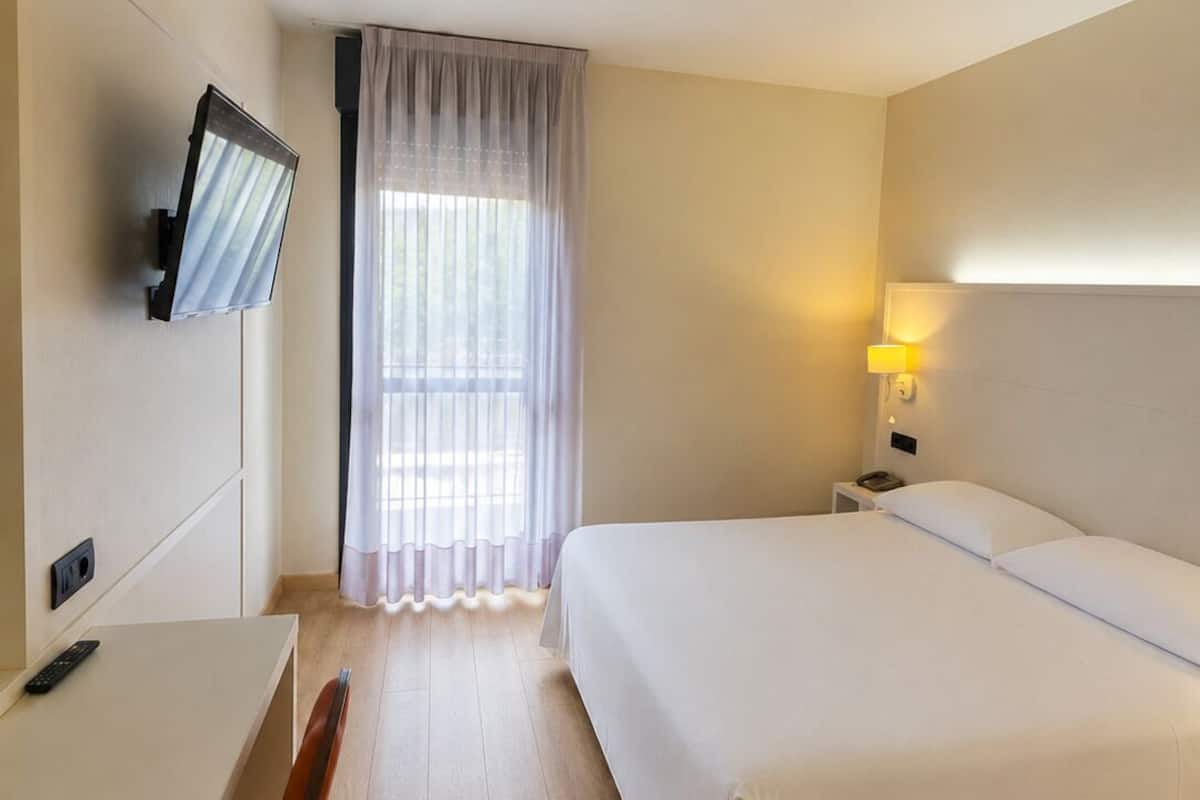 A hotel room with a bed, a desk, a chair, a TV, a lamp, and a window with curtains.