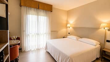 Een hotelkamer met een bed, bedlampen, een bureau met twee wijnglazen, een stoel en een raam met dunne gordijnen.