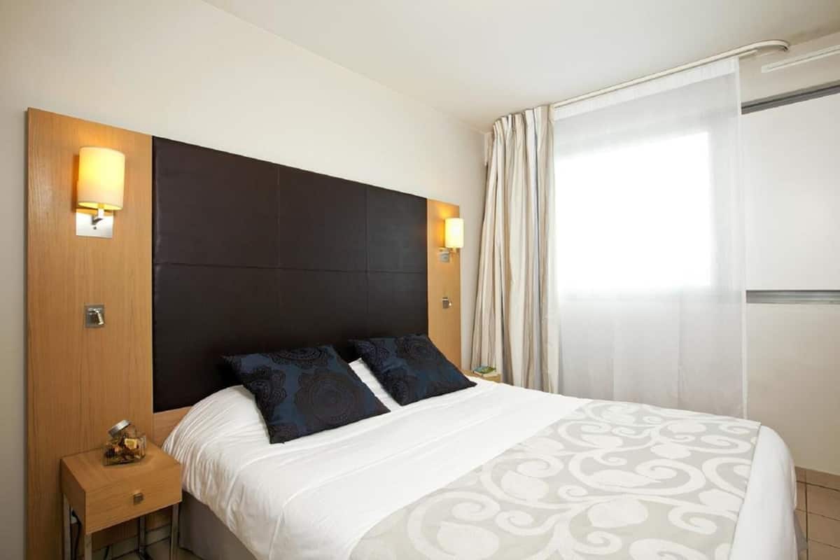 Una habitación de hotel con cama, lámparas de noche, mesita de noche y ventana con cortinas.