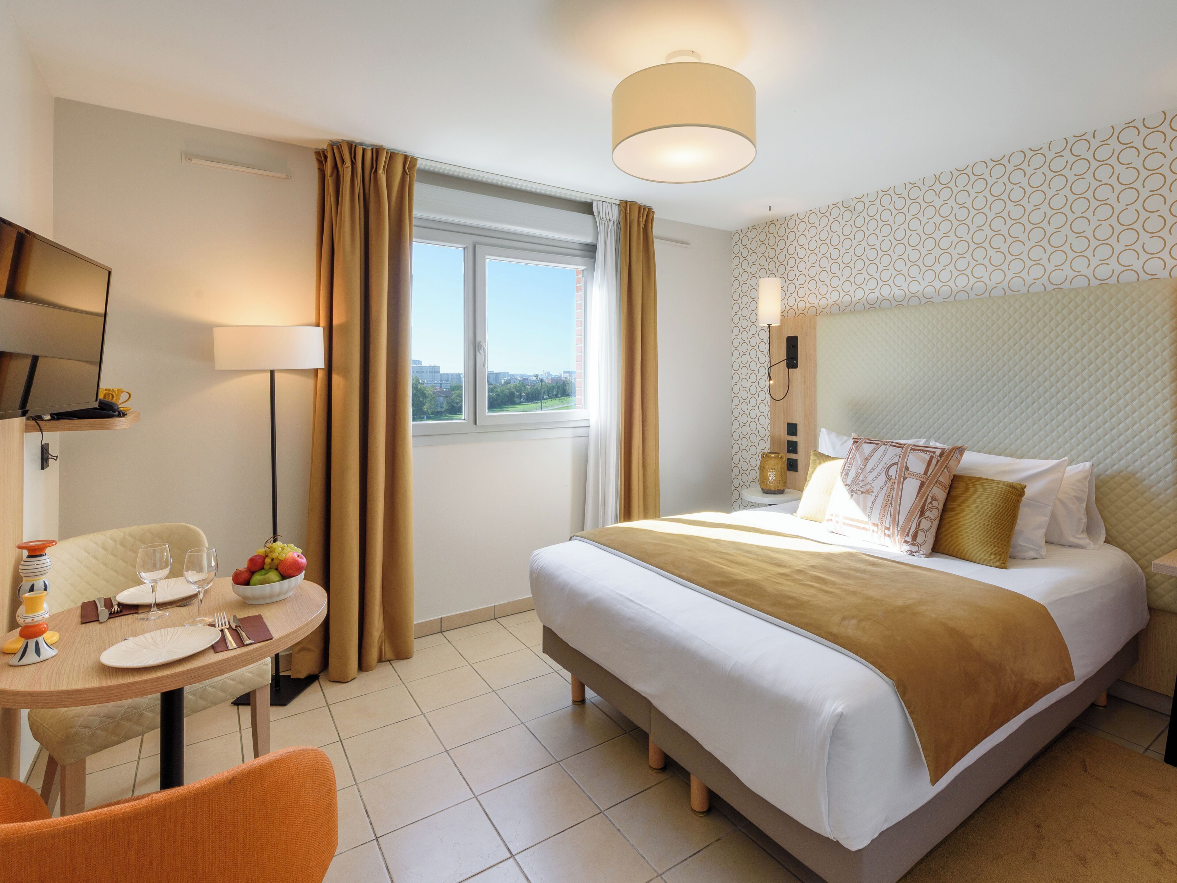 A hotel room with a bed, a desk with a chair, a television, and a window with curtains.