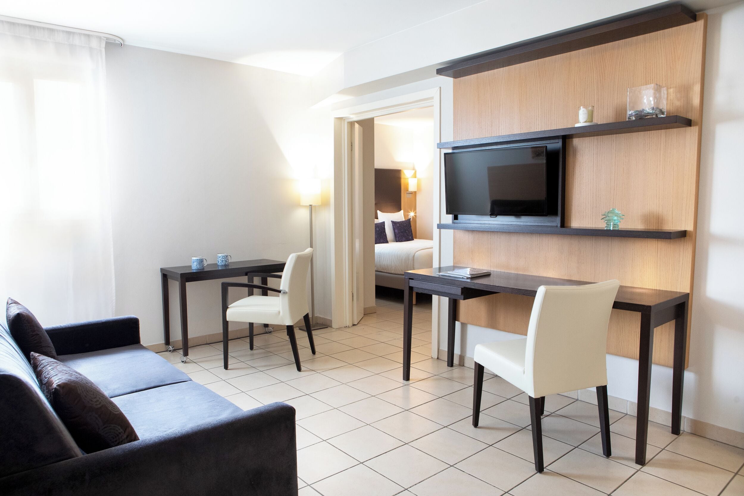 A modern hotel room with a sofa, a dining table with chairs, a TV mounted on a wooden panel, and a bed with a nightstand.