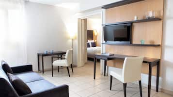 A modern hotel room with a sofa, a dining table with chairs, a TV mounted on a wooden panel, and a bed with a nightstand.