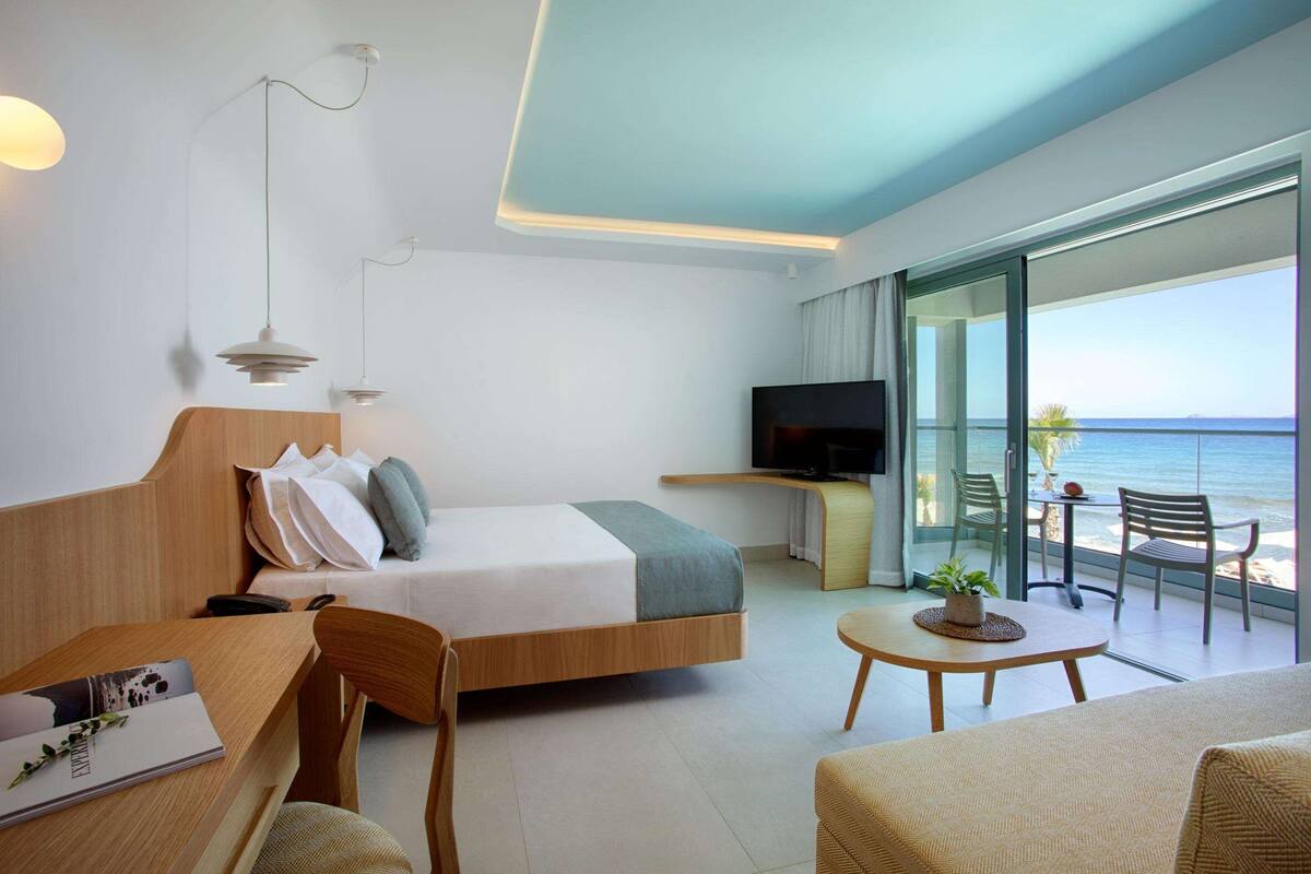 A modern hotel room with a bed, a desk, a TV, and a balcony with a view of the ocean.