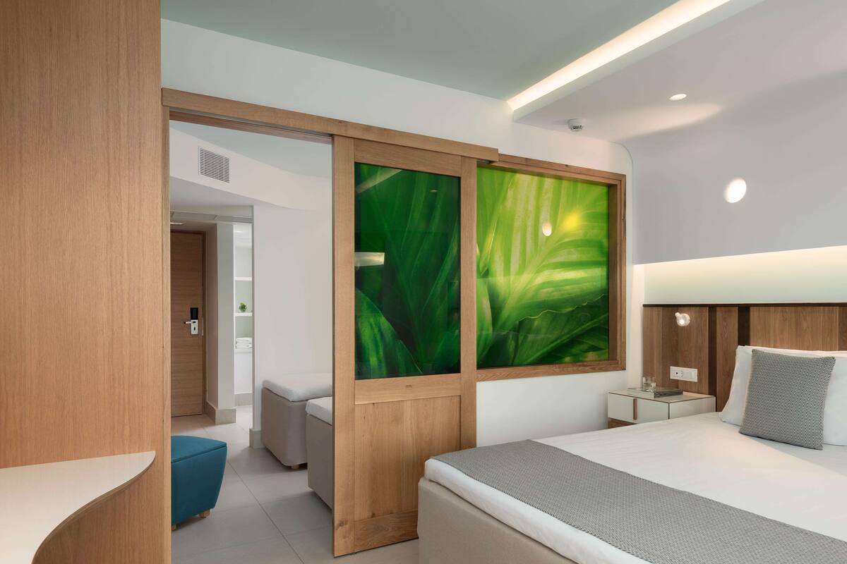 A modern hotel room with a bed, a nightstand, a desk, a blue chair, and a sliding door with a green leaf pattern.