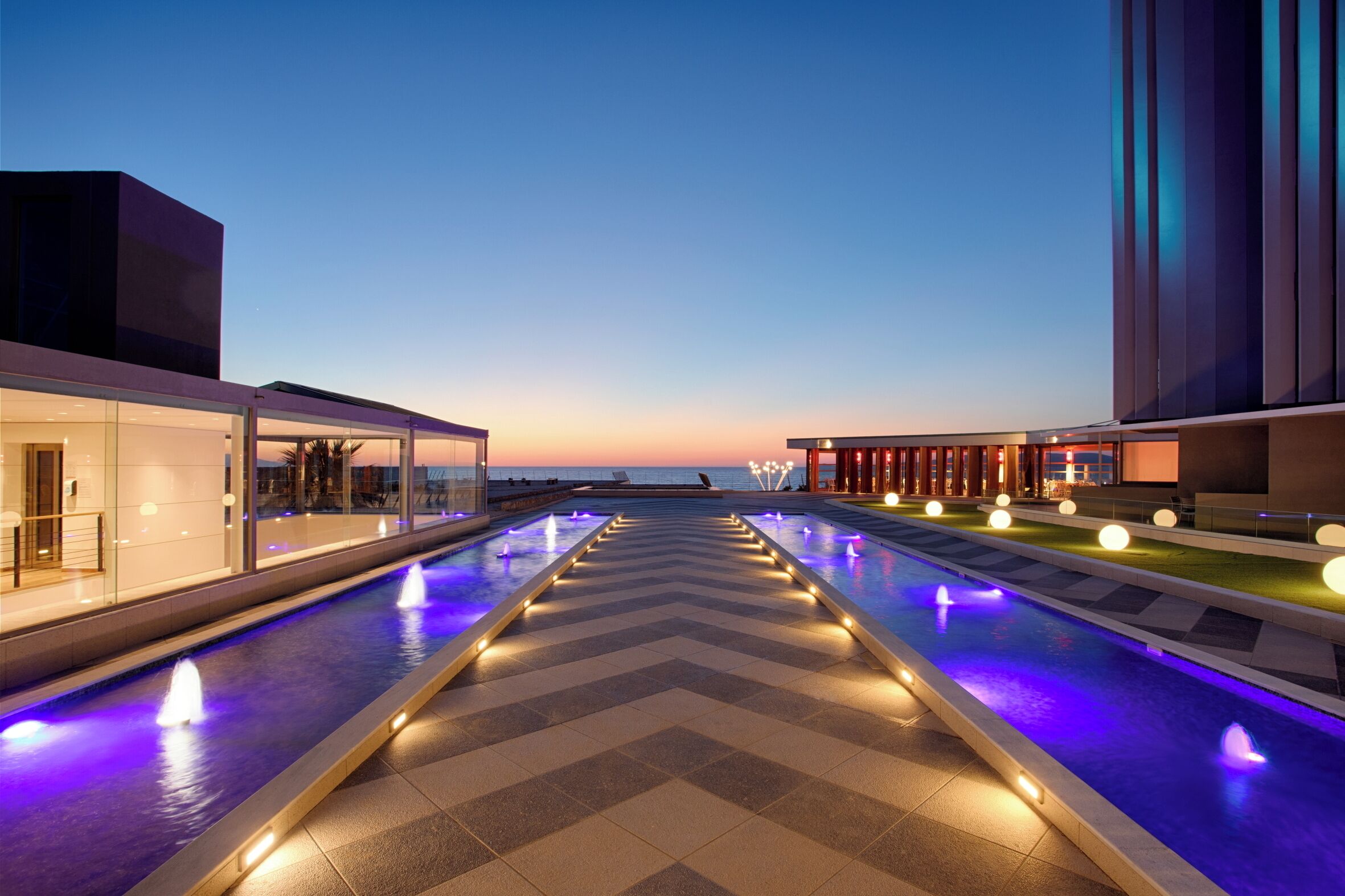 Un espace piscine moderne dans un hôtel, avec un motif en chevron, des jeux d’eau et une vue sur la mer au crépuscule.